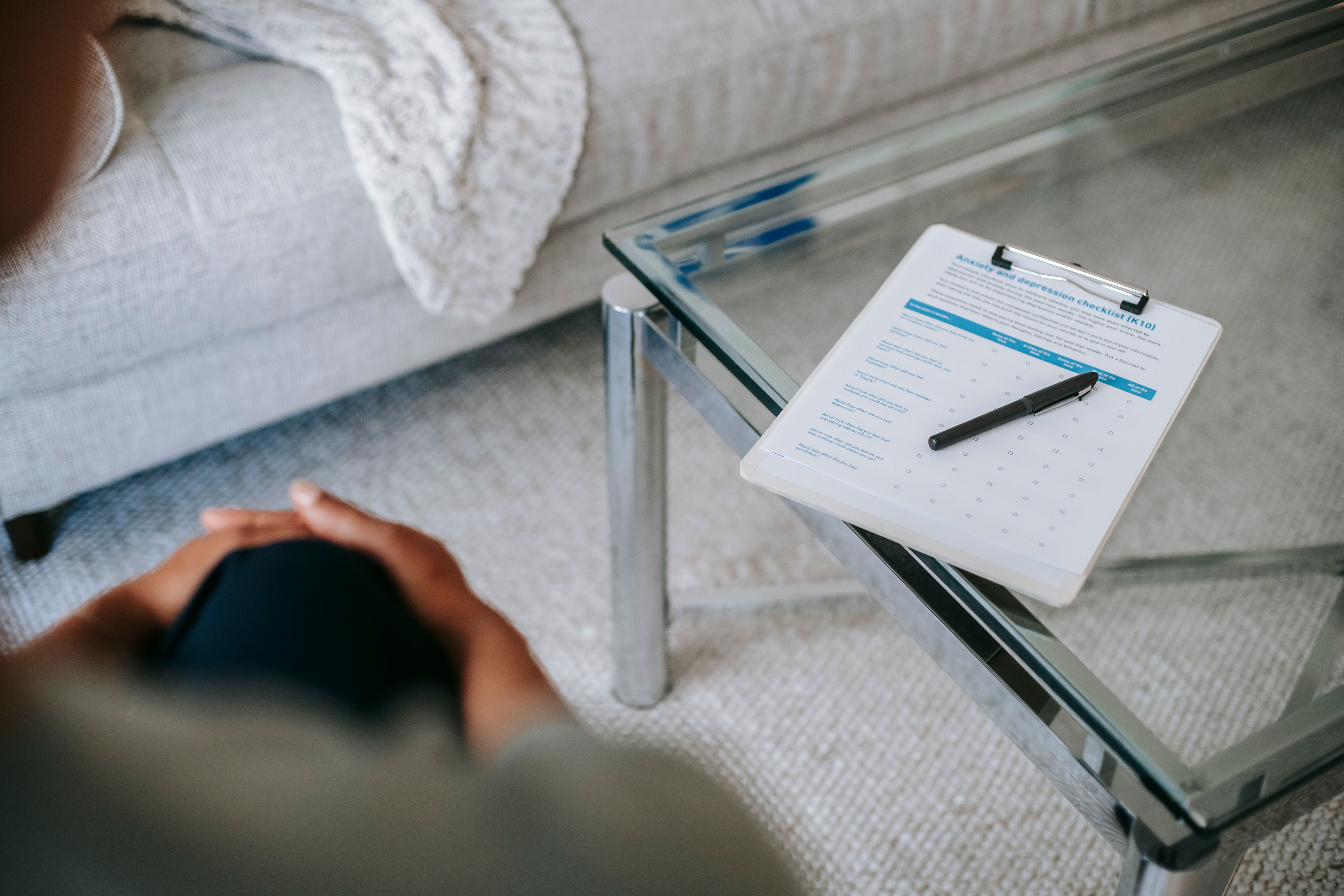 A clipboard holding a checklist document resting on a glass coffee table in front of a modern living area sofa.