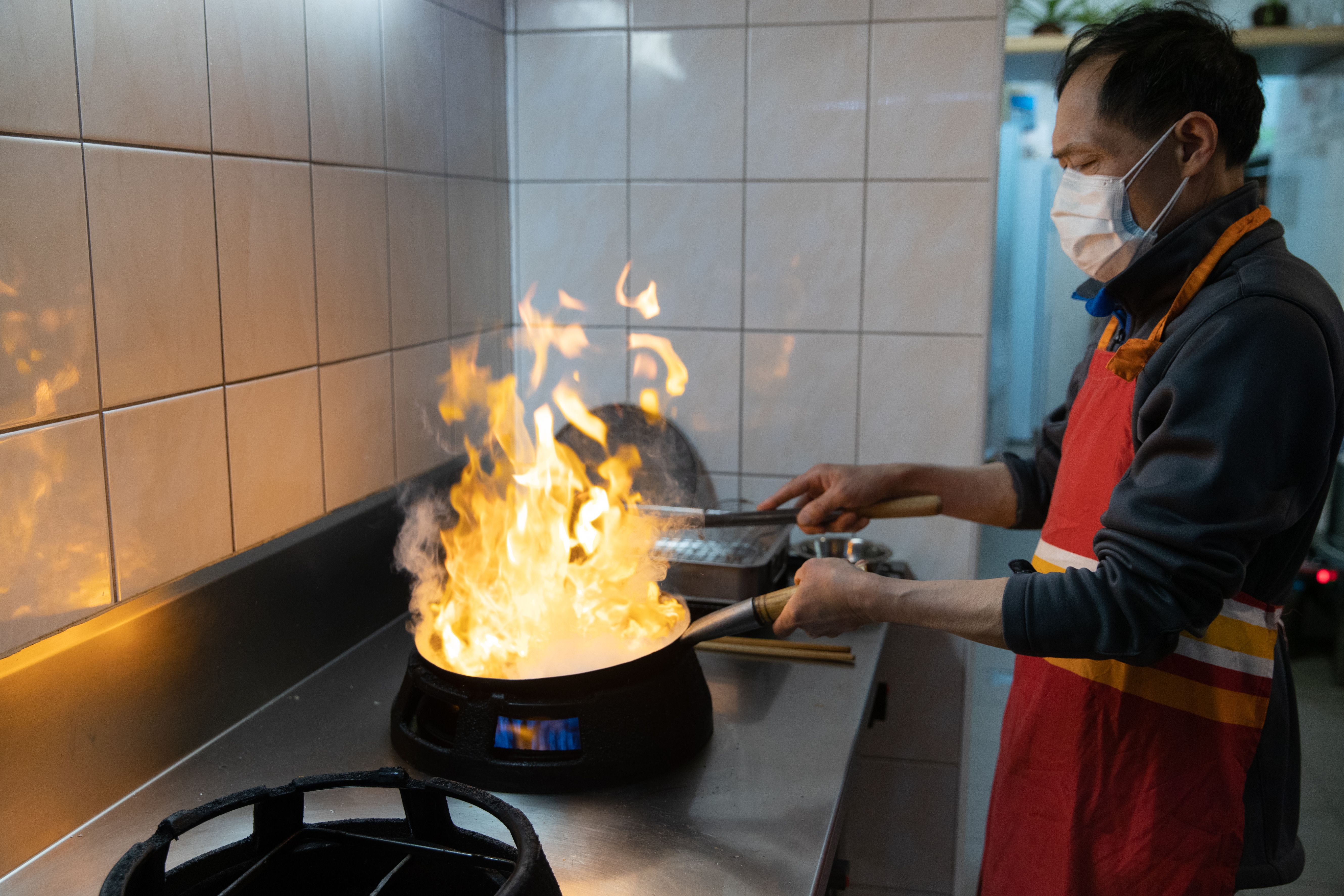 A cook wearing a protective face mask and red apron managing high flames while stir-frying in a wok over a gas stove.