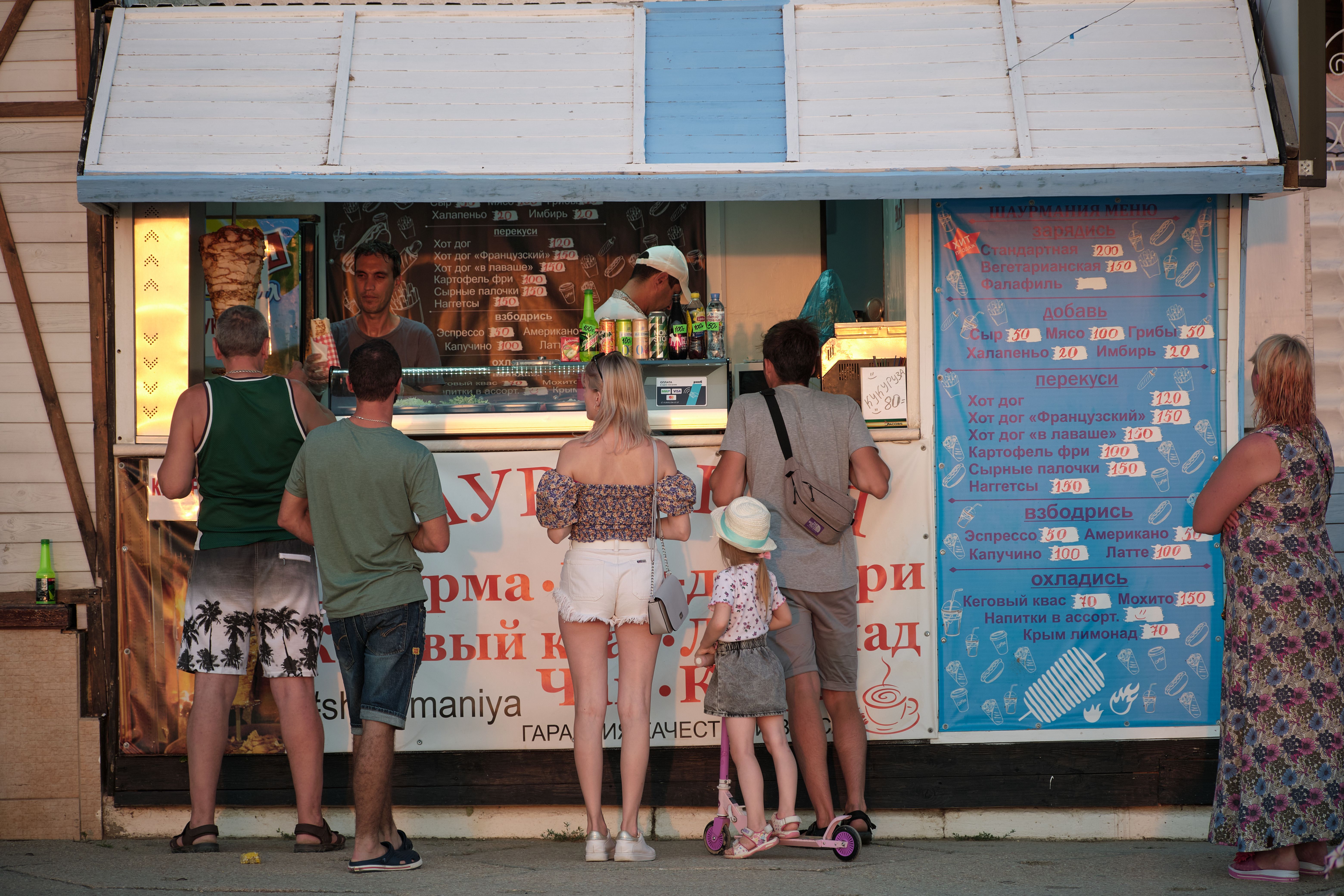 A line of customers waiting at an outdoor food kiosk stand displaying a menu board in Russian.