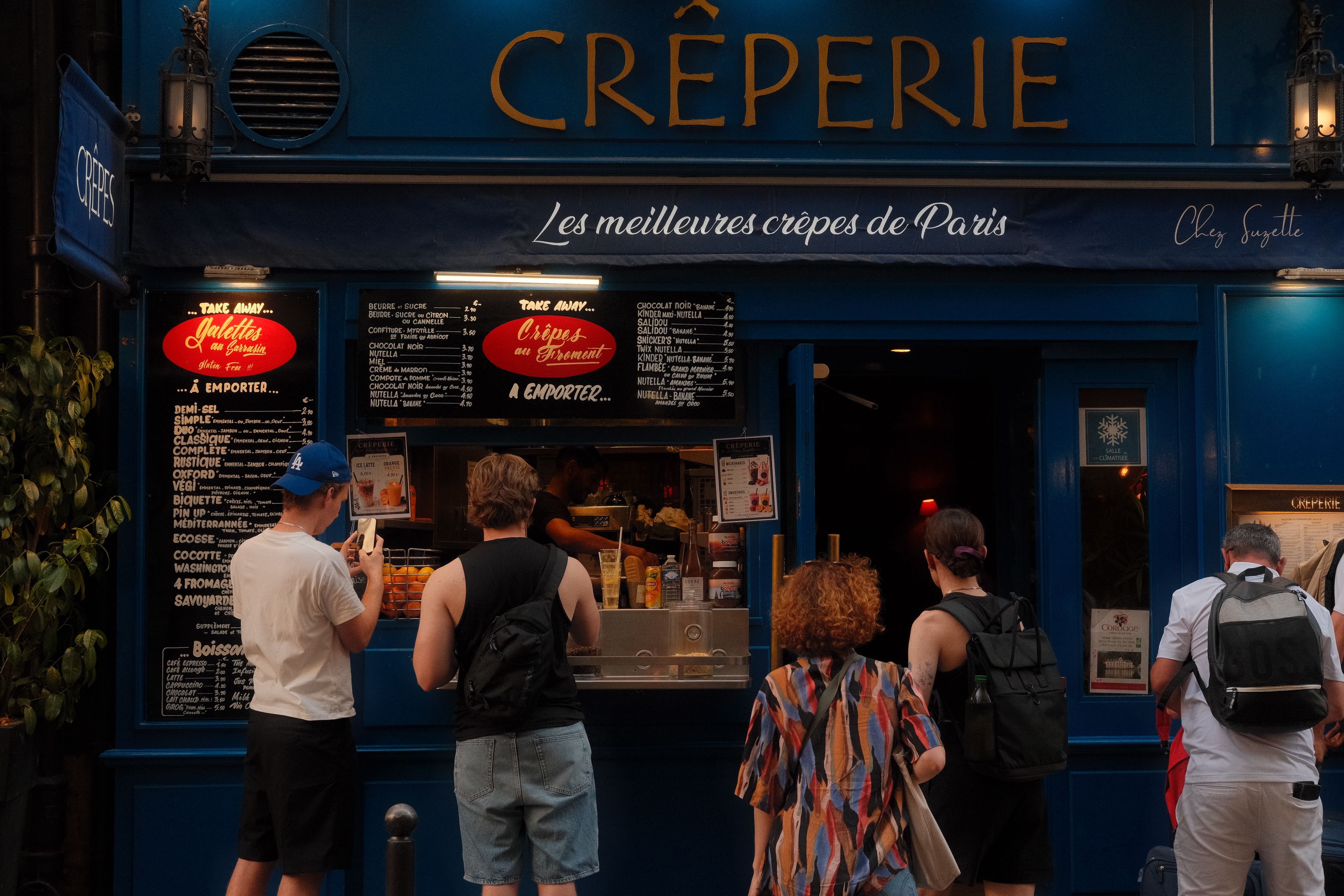 People standing outside a blue-fronted French crêperie storefront in Paris reading takeaway menu boards.