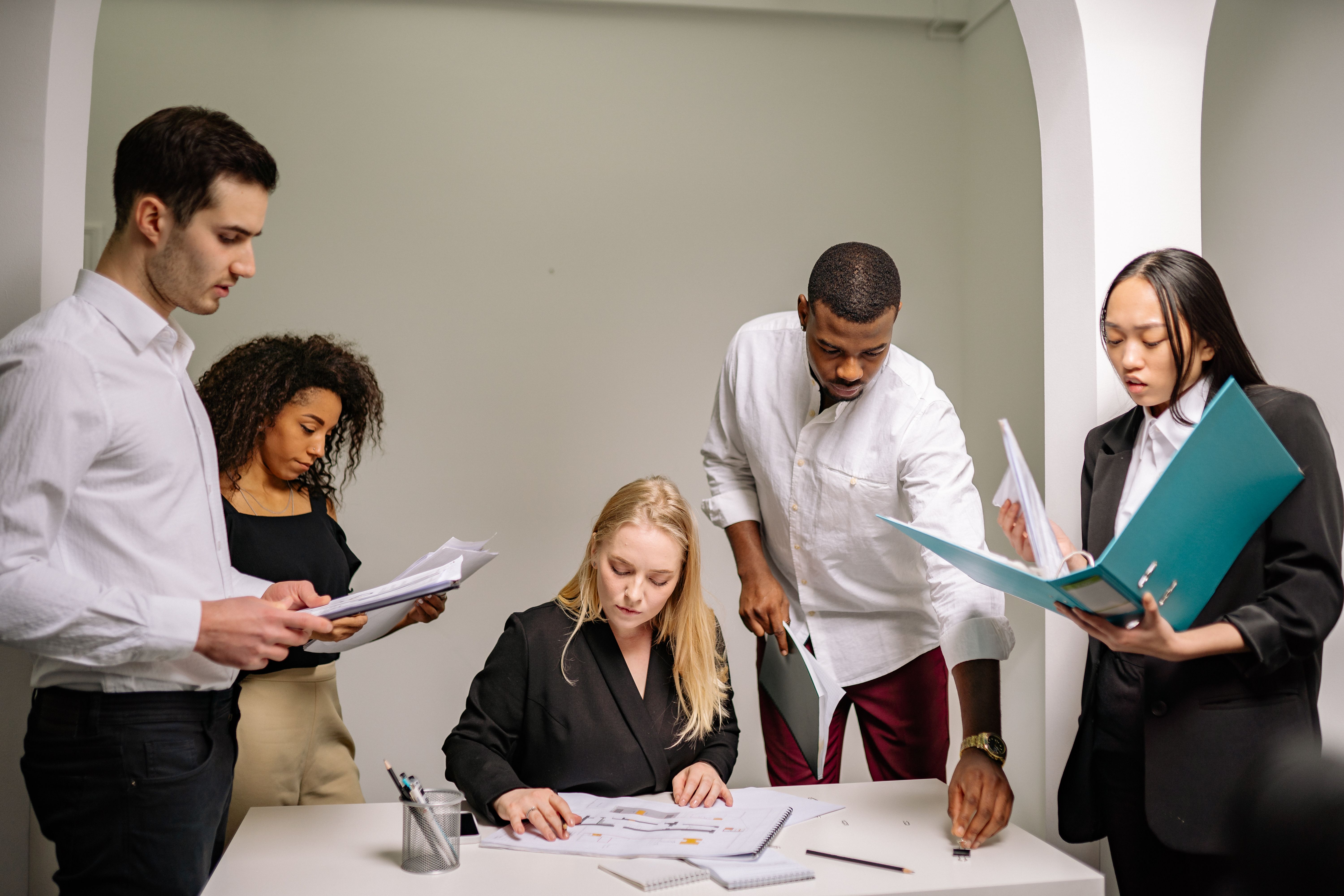 A group of five professionals gathered around an office desk reviewing folders, documents, and architectural plans.
