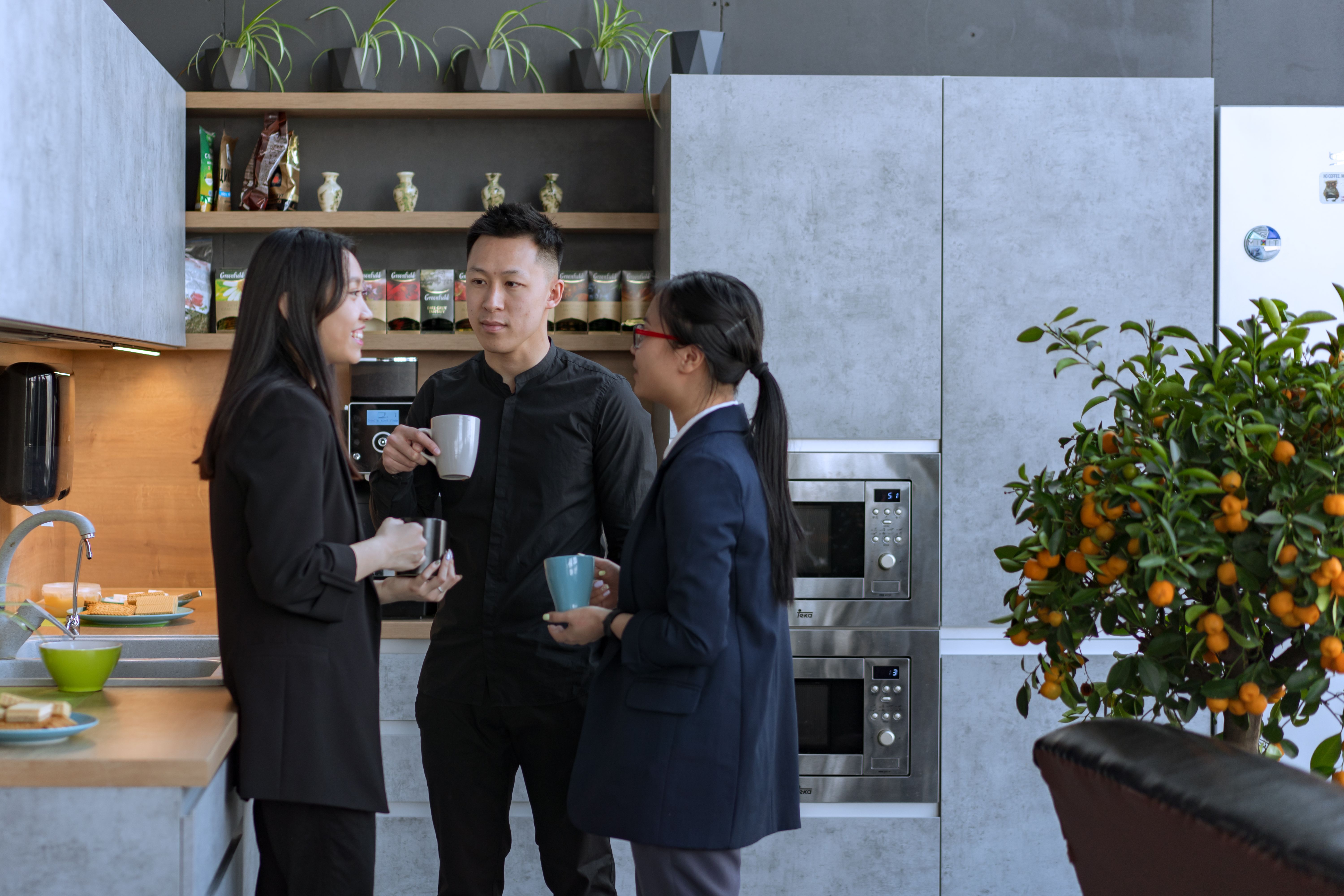 Three colleagues standing in a modern office kitchen area holding mugs and chatting near a potted citrus plant.