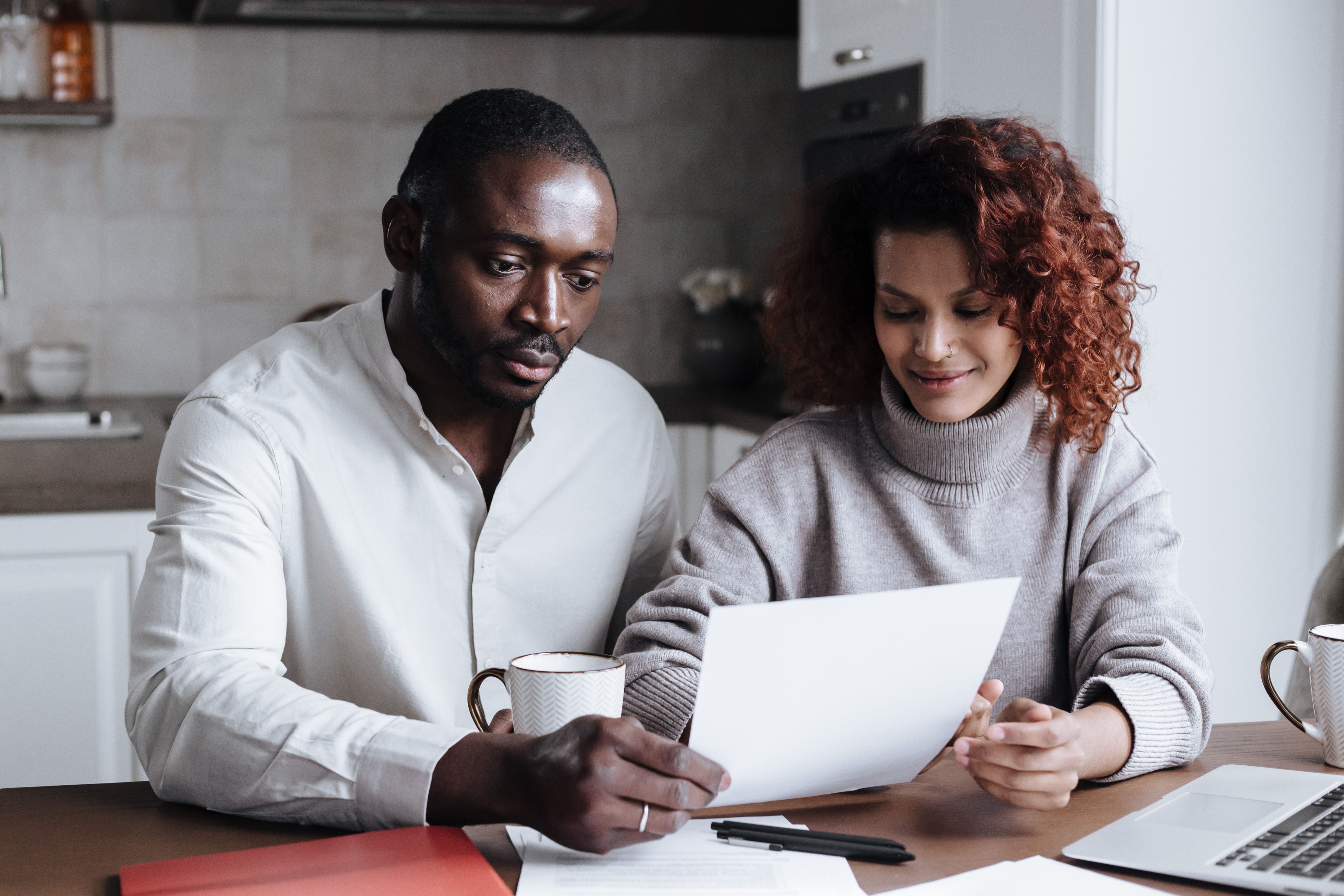 A man and a woman sitting together at a table reviewing paperwork with a laptop and coffee mug nearby.