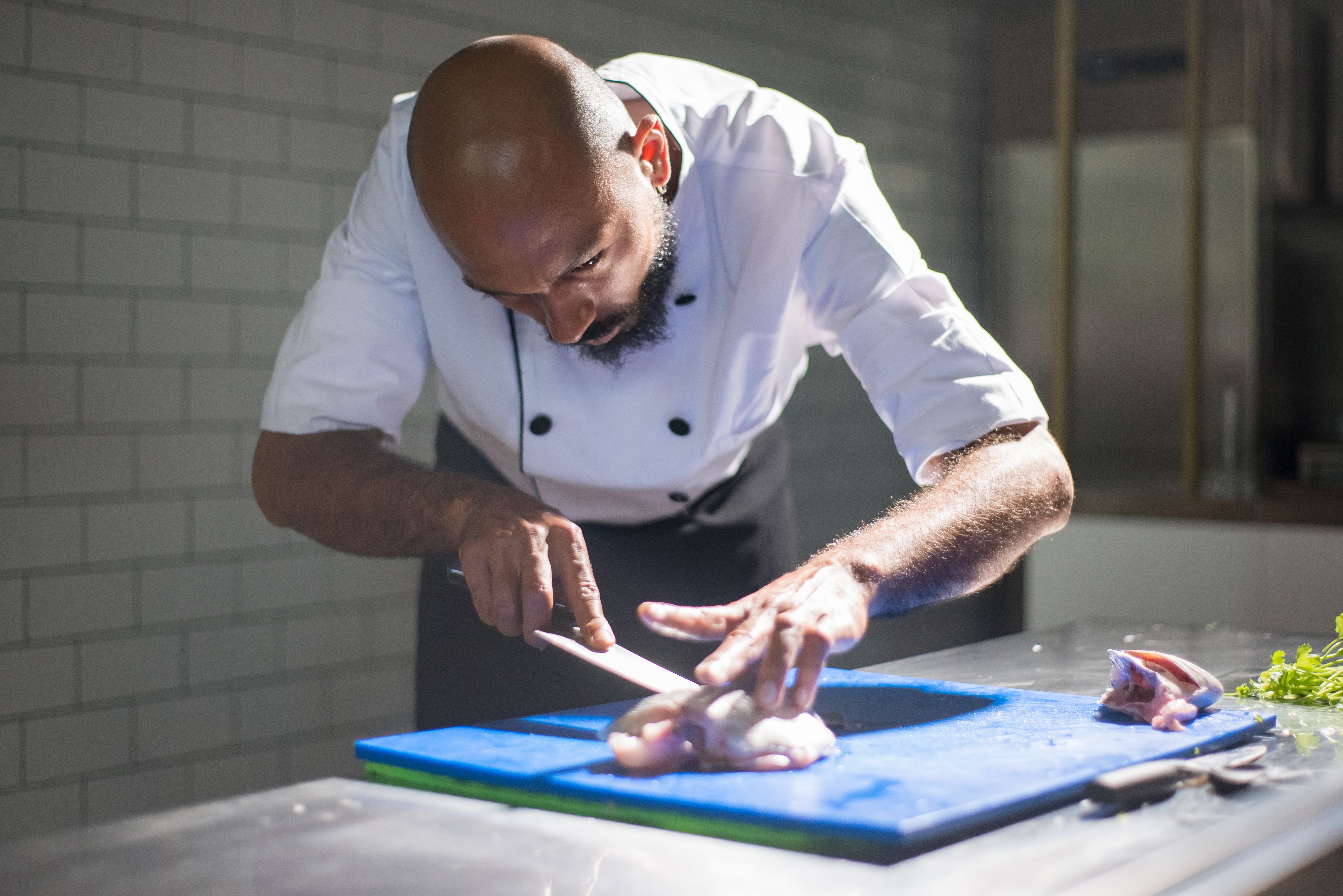 A chef in a white jacket carefully slicing raw fish on a blue cutting board in a commercial kitchen setup.