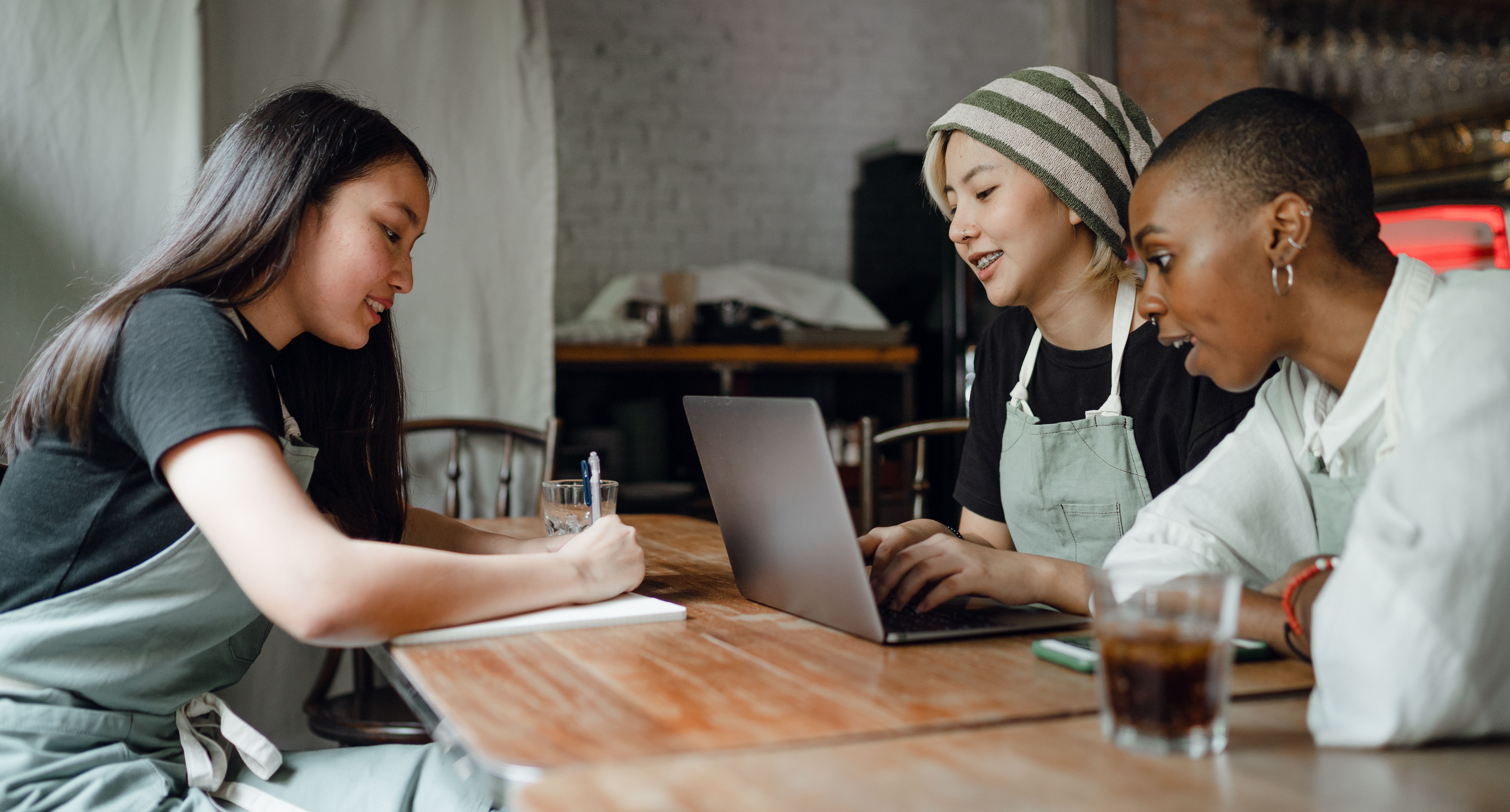 Three young cafe colleagues sitting at a wooden table together working on a laptop and taking notes in a notebook.