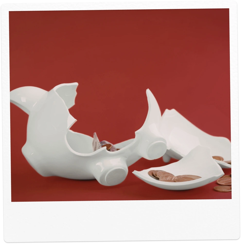 Casse-tirelire en forme de cochon blanc brisé avec des pièces de monnaie dispersées autour sur fond rouge.