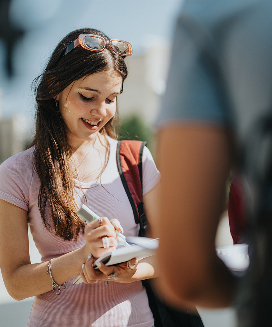 Lächelnde Jugendliche mit langen dunklen Haaren schreibt in ein Notizbuch im Freien