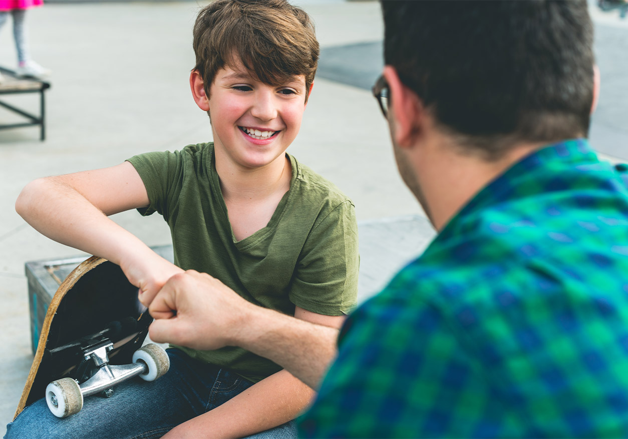 Lächelnder Junge mit Skateboard gibt einem erwachsenen Mann mit Brille einen Faustgruß auf einem Skatepark-Platz