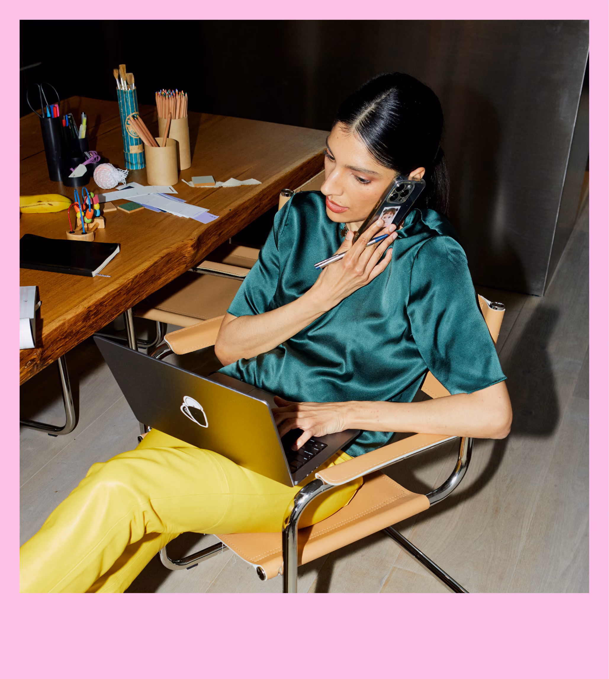 Woman in yellow pants and green blouse talking on phone while typing on laptop in office chair.