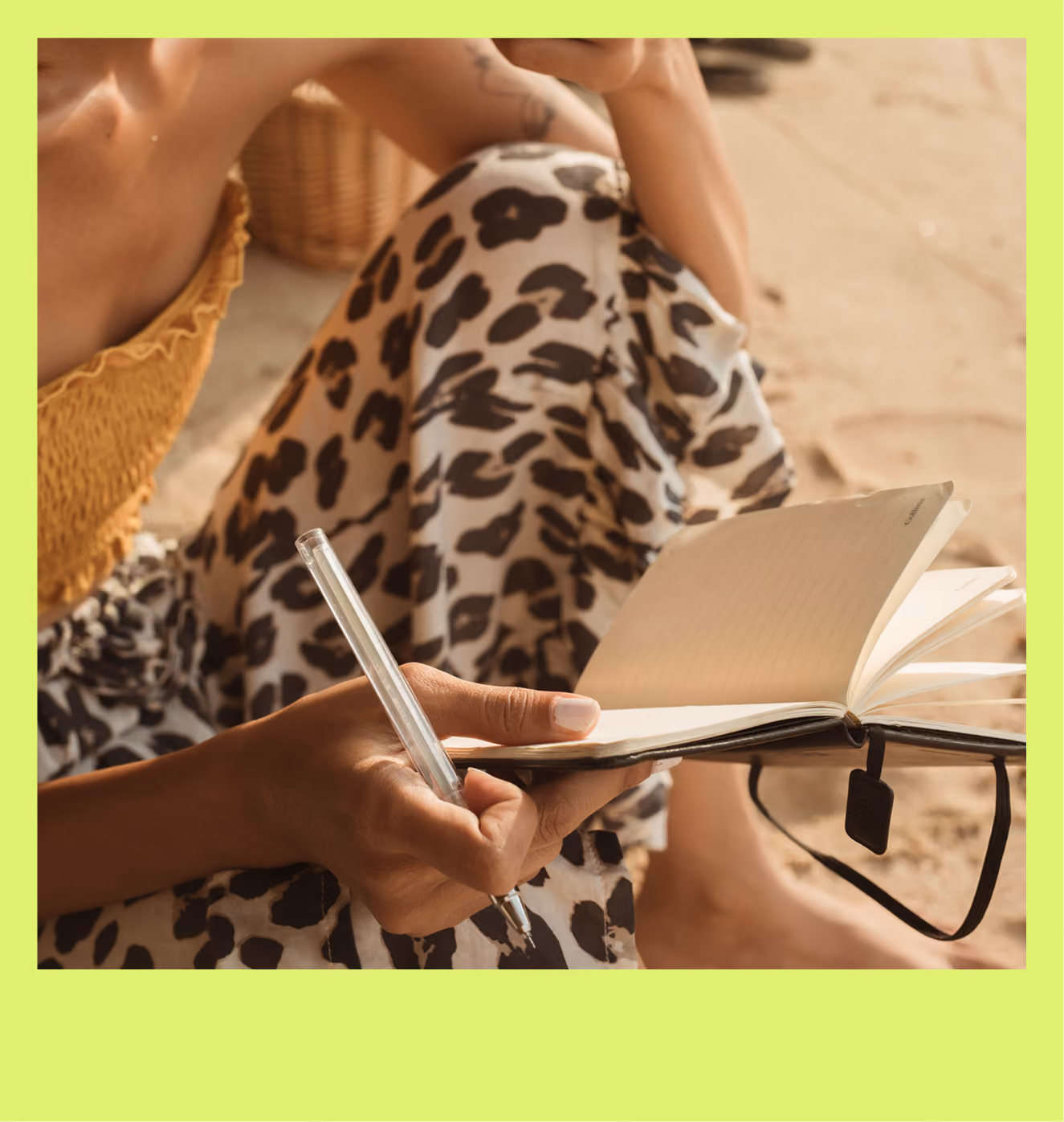 Person sitting on sand in leopard print pants, writing in a notebook with a clear pen.
