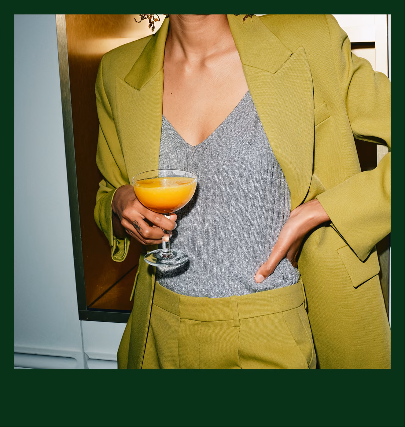 Person in olive green suit and silver top holding a cocktail glass with orange drink.