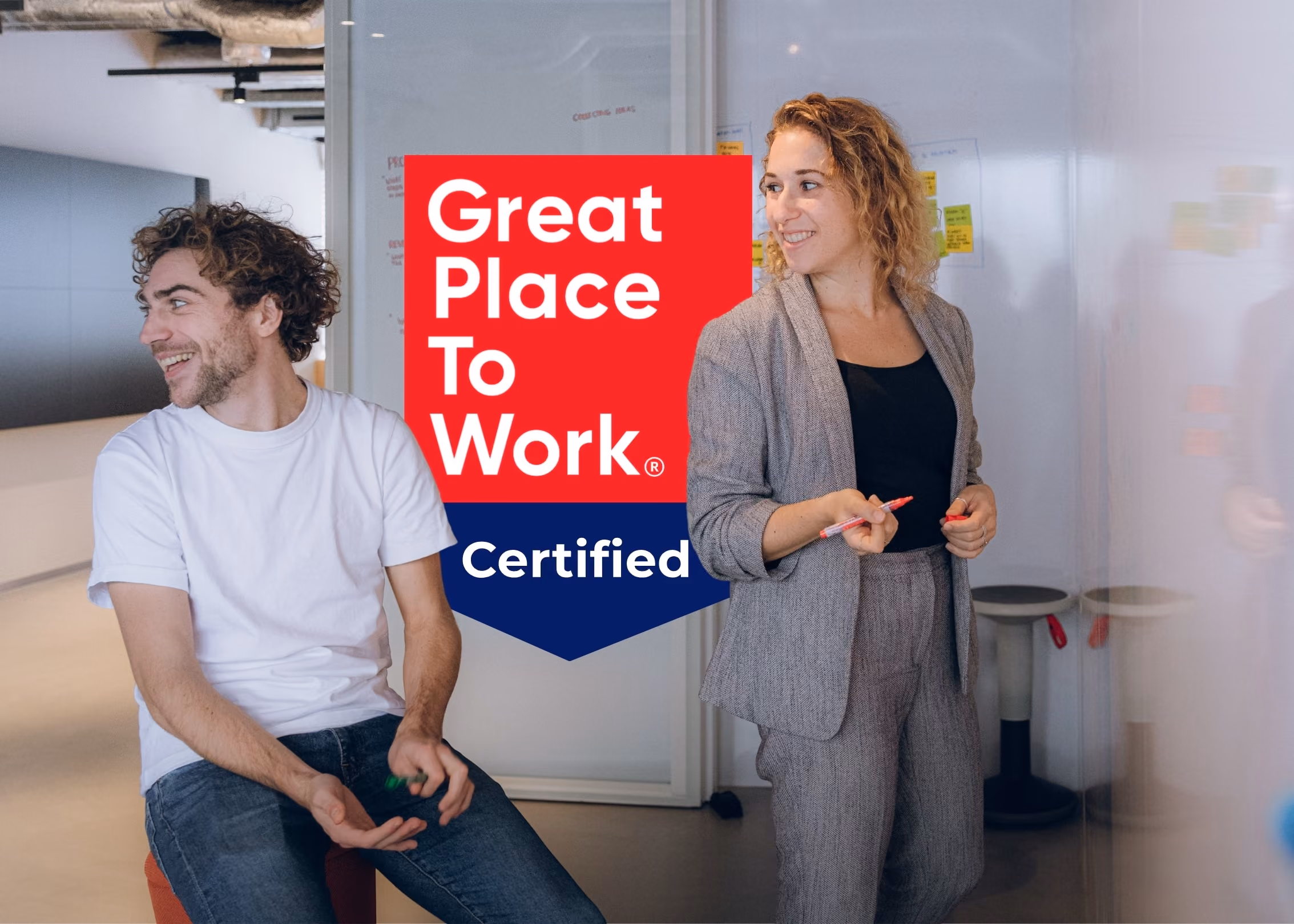 Gruppe på fem personer til et uformelt møde på et farverigt, moderne kontor med etageopdelte siddepladser og et "Great Place to Work"-certificeringsmærke lagt henover.