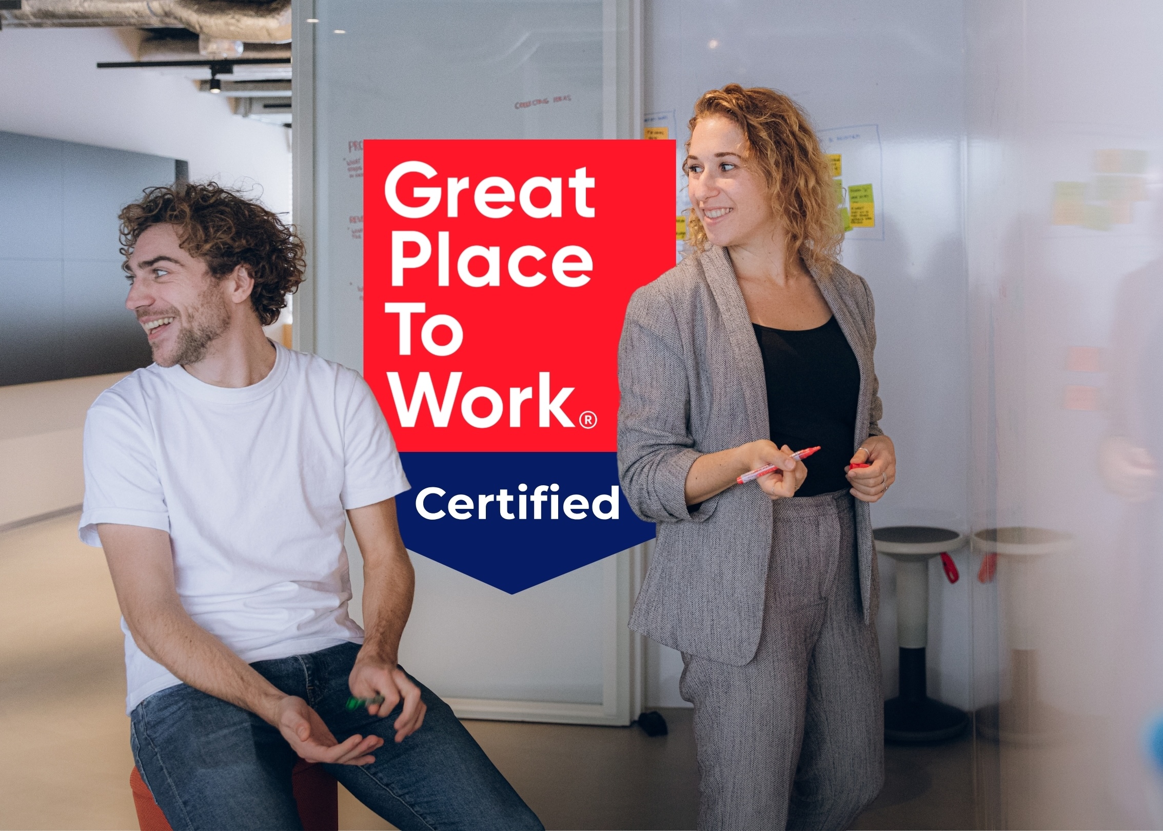 Group of five people having a casual meeting in a colorful modern office with tiered seating and a Great Place to Work certification badge overlay.