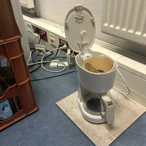 Alte Kaffeemaschine mit offenem Deckel und Filterpapier auf einem Bürotisch
