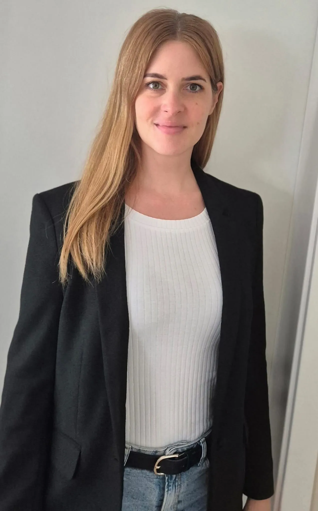 Frau mit langen blonden Haaren trägt schwarzen Blazer, weißes geripptes Oberteil und Jeans mit schwarzem Gürtel.
