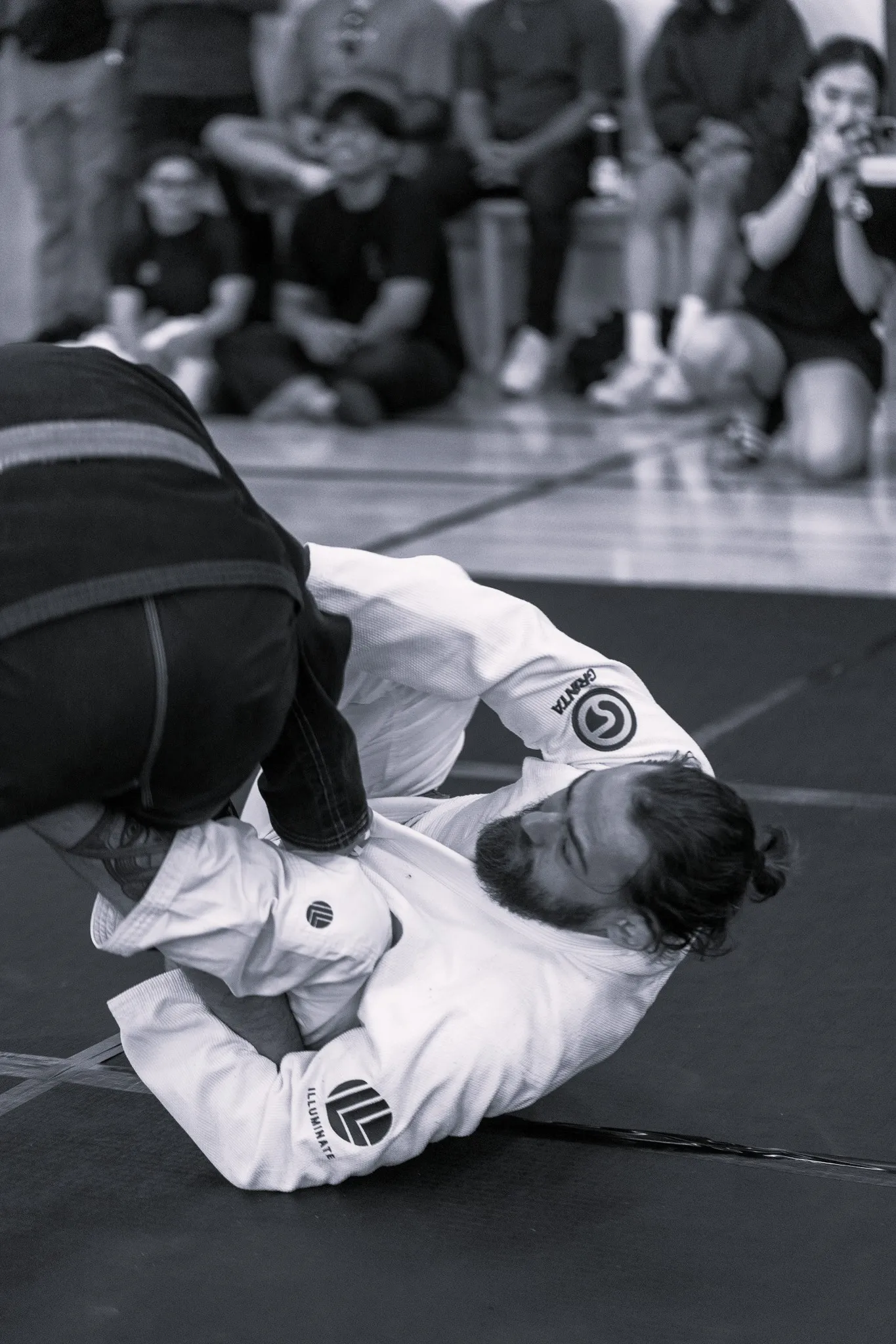 Two men grappling in Brazilian Jiu-Jitsu match with one wearing a white gi and the other in black, while spectators watch.
