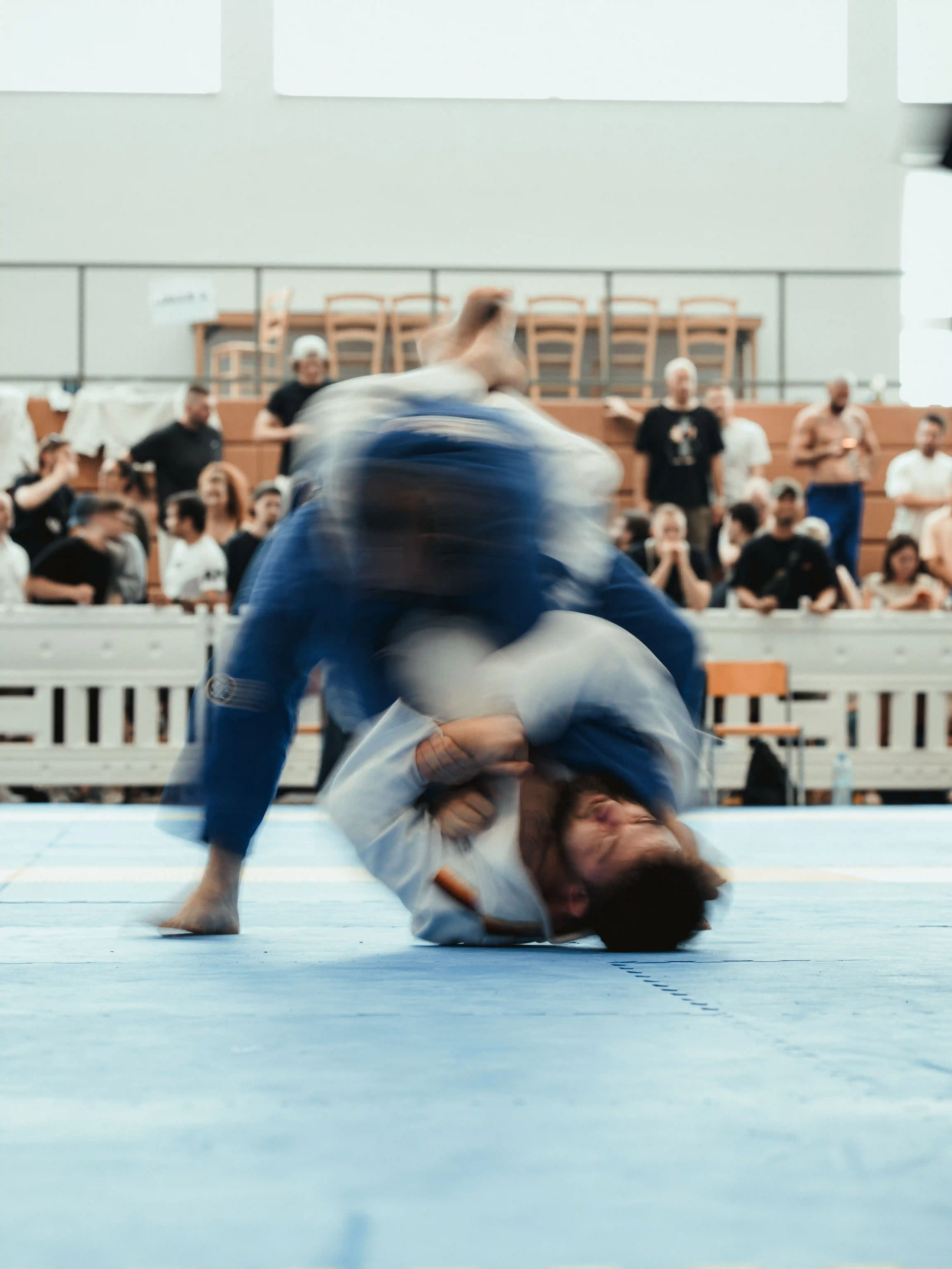 Two men grappling in Brazilian Jiu-Jitsu match with one wearing a white gi and the other in black, while spectators watch.