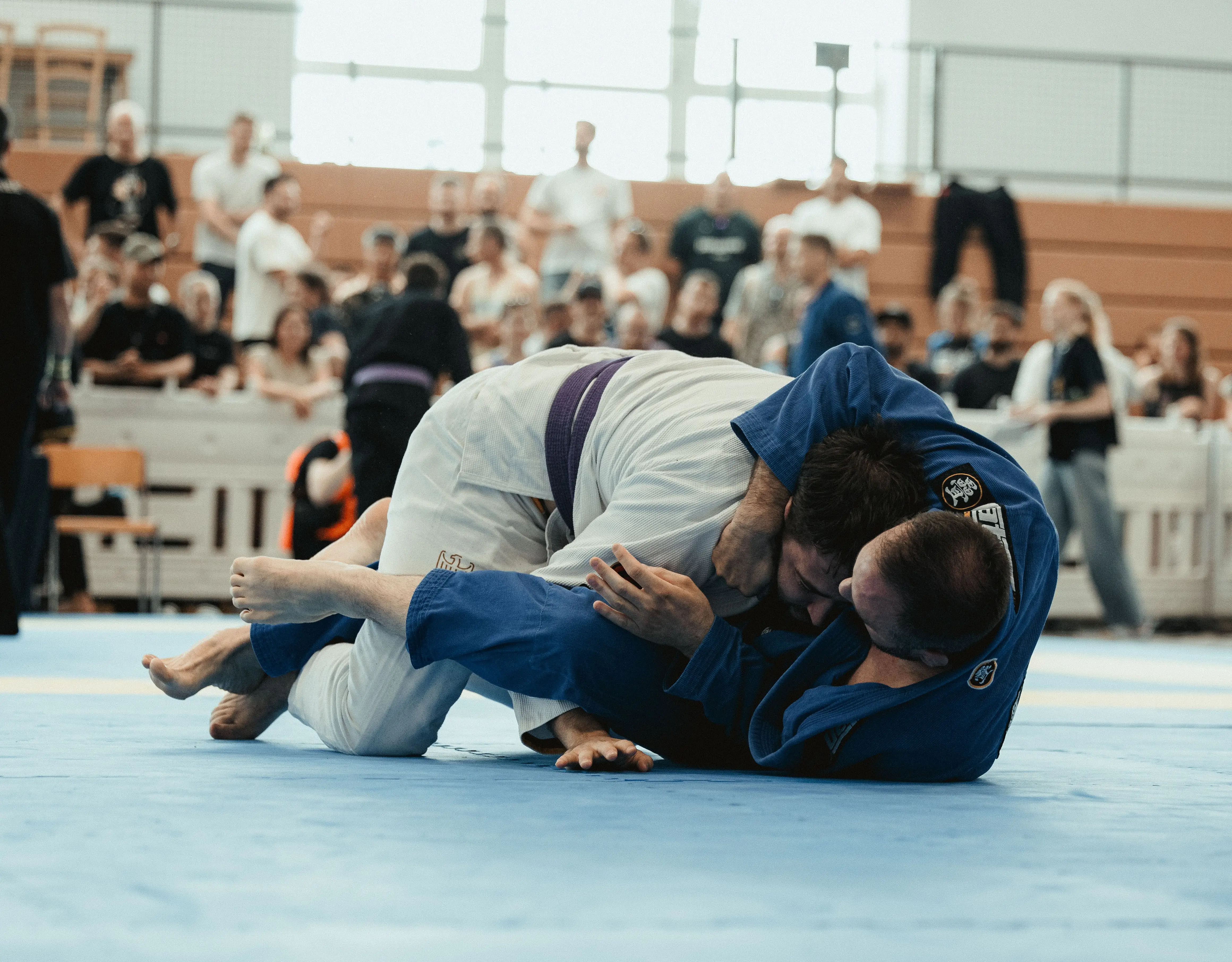 Zwei Männer im Brazilian Jiu-Jitsu kämpfen auf blauer Matte vor Zuschauern in einer Halle.
