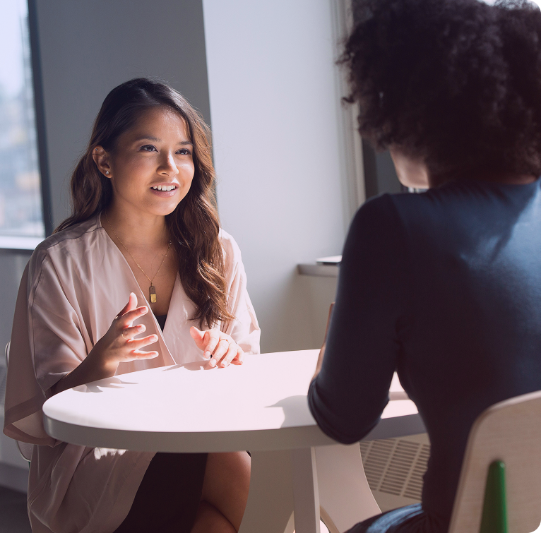 Two women sitting at a round table engaged in a conversation, one speaking with expressive hand gestures.