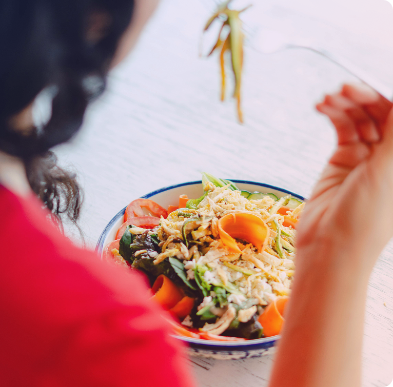Person in a red shirt eating a colorful salad with tomatoes, lettuce, shredded cheese, and carrot ribbons using chopsticks.