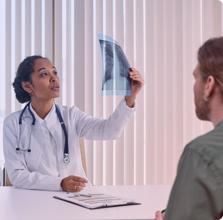 Female doctor with stethoscope examining a chest X-ray while consulting a male patient at a desk.