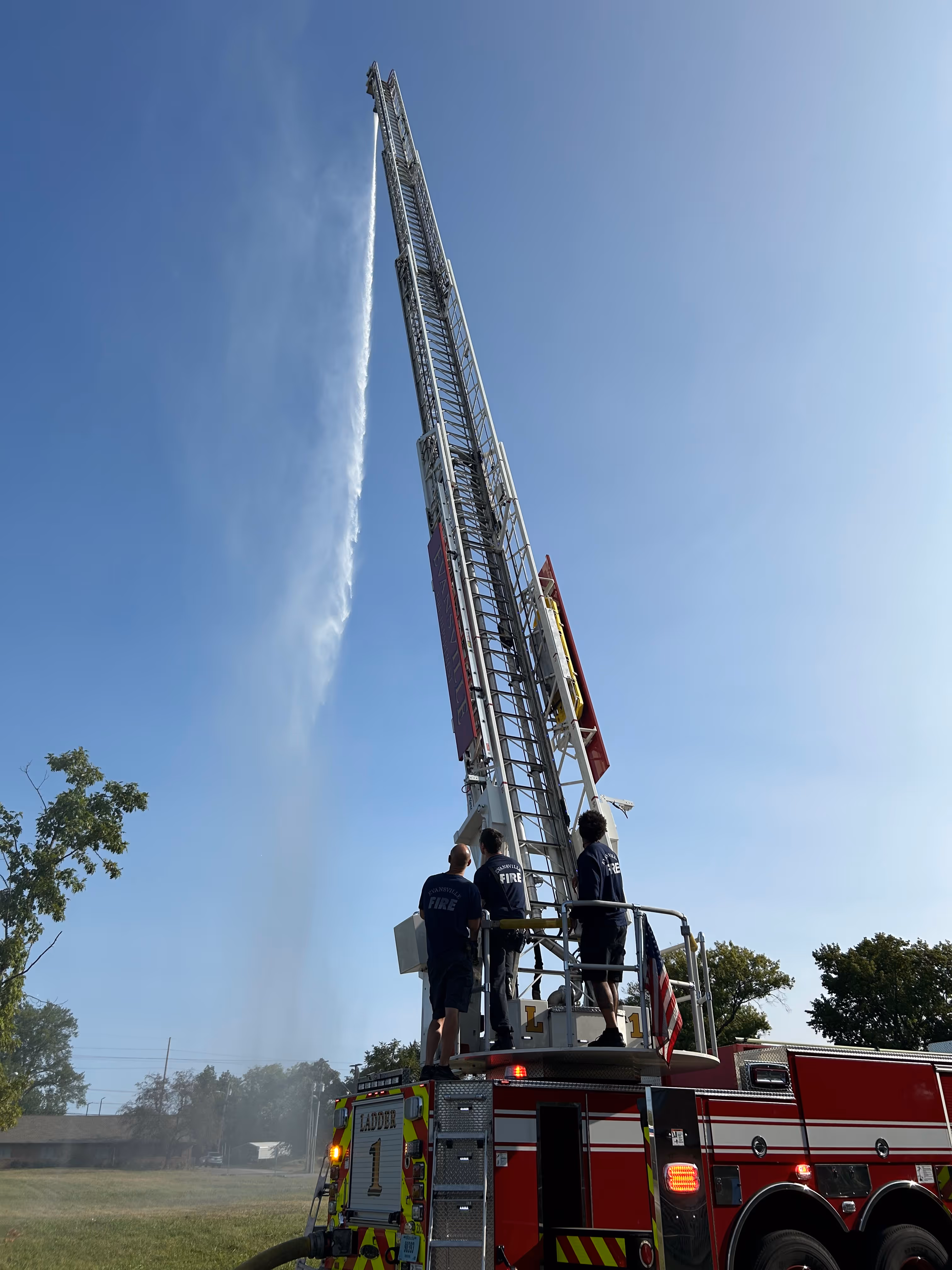 Three firefighters standing on an elevated platform of a red fire truck spraying water high into the clear blue sky.
