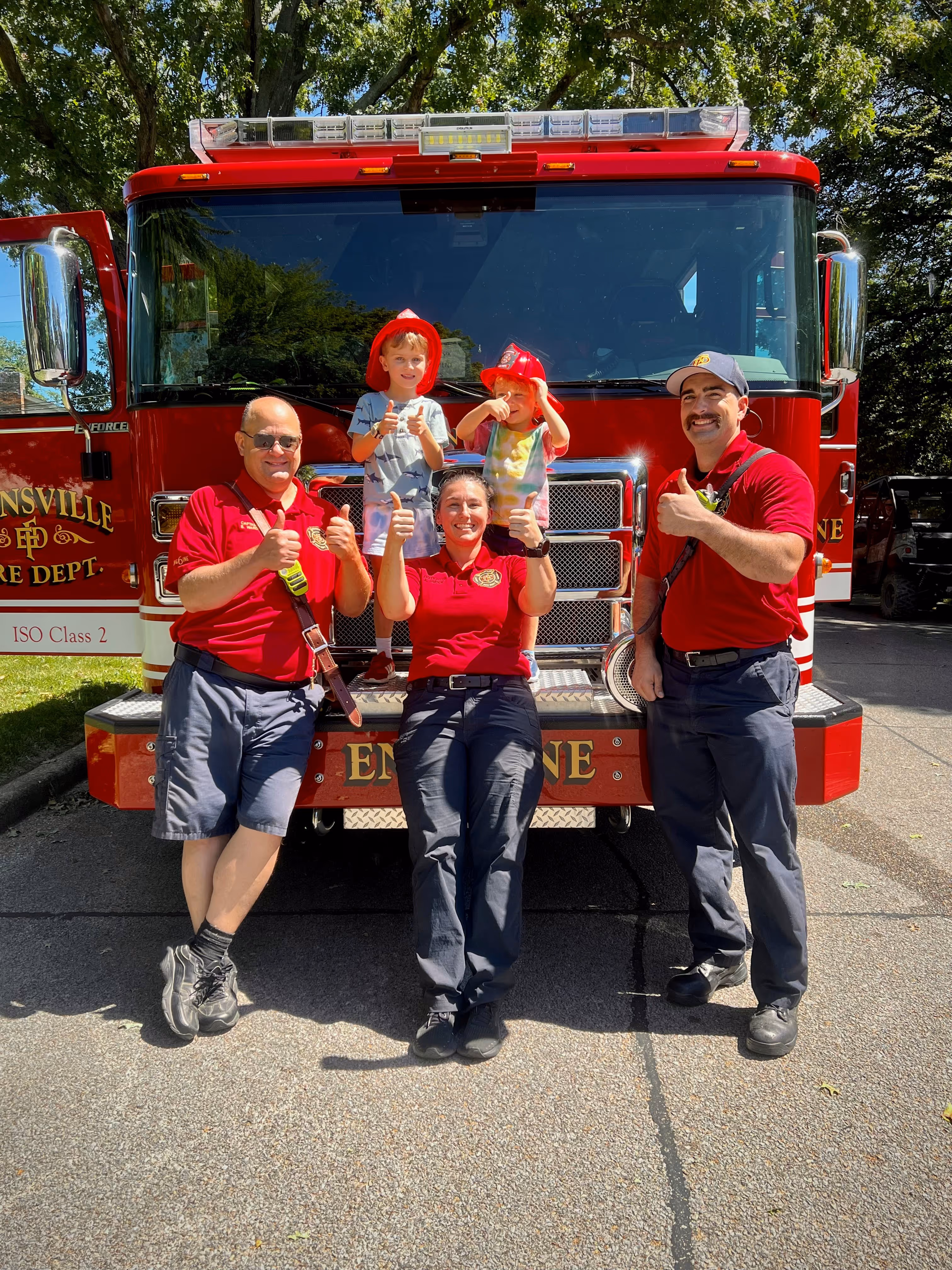 Three firefighters in red shirts and two children wearing red firefighter hats pose with thumbs up in front of a red fire truck.