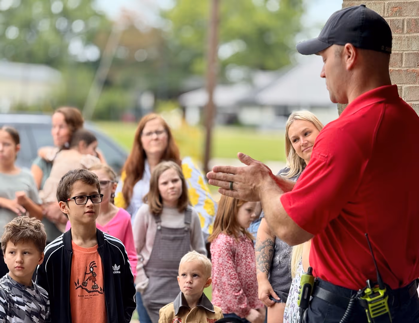 Man in red shirt speaking to a group of attentive children and adults outdoors.