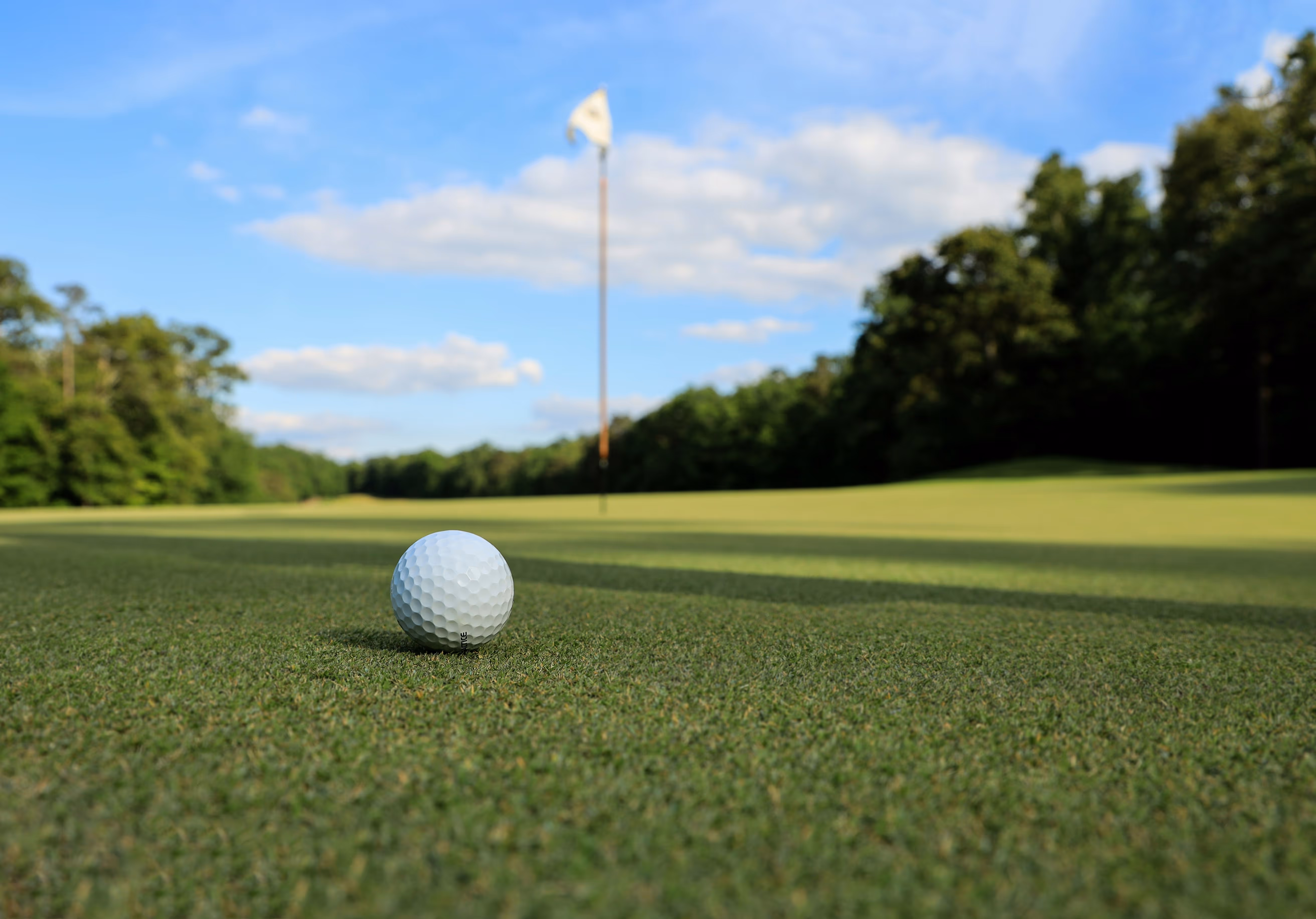 Close-up of a white golf ball on green grass with a blurred golf hole flag in the background under a blue sky.