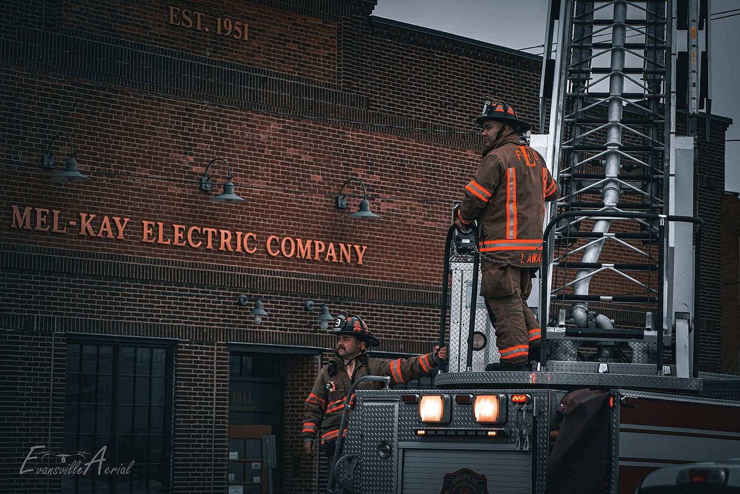 Two Evansville firefighters in turnout gear stand on a ladder truck outside the Mel-Kay Electric Company building during a fire response.