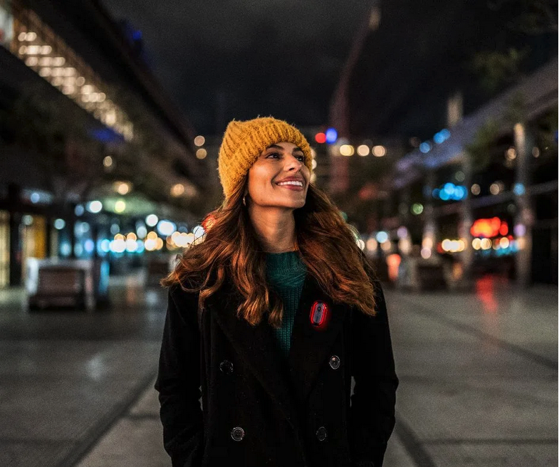 Young man with glasses and wavy hair wearing a black sweater sitting next to a beige wall with dried plants.