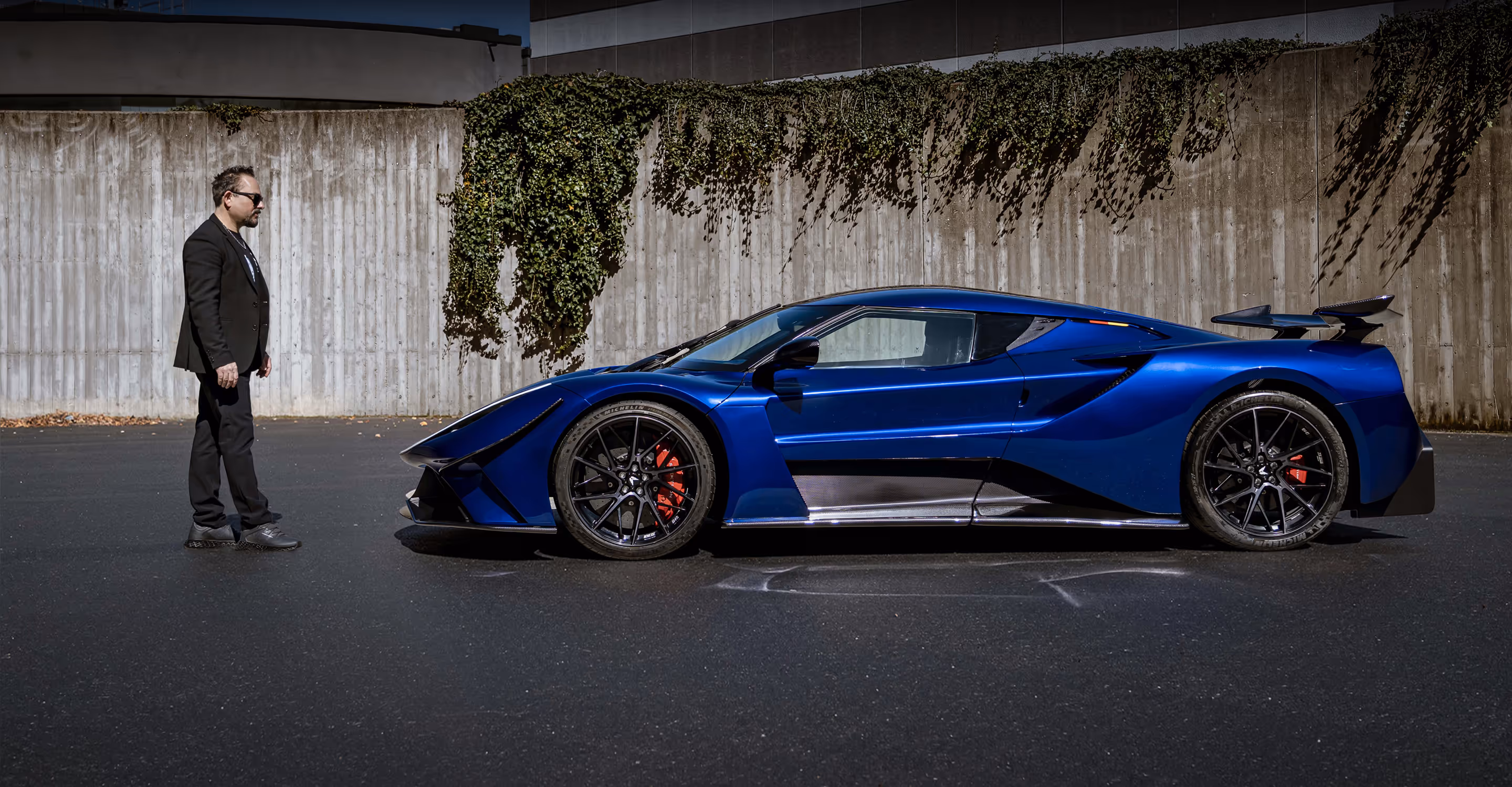 Silhouetted man in profile standing in a dimly lit garage with a blurred sleek blue sports car in the background.