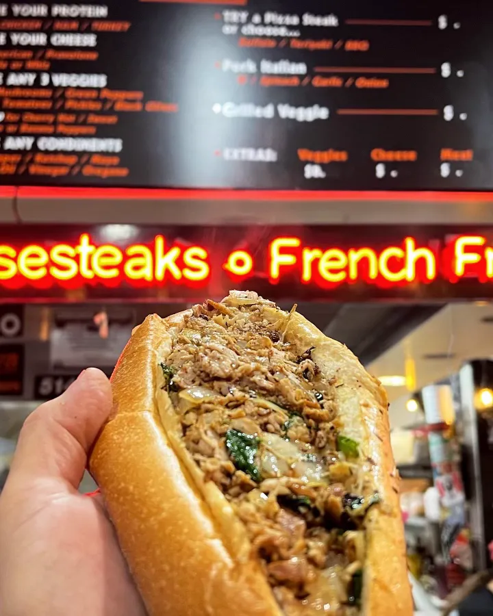 Hand holding a Philly cheesesteak sandwich filled with cooked meat and vegetables in front of a neon sign reading 'Cheesesteaks o French Fr...'.
