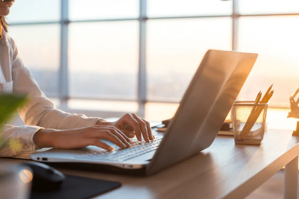 Person typing on a laptop keyboard at a wooden desk with pencils in a holder and sunlight coming through large windows.