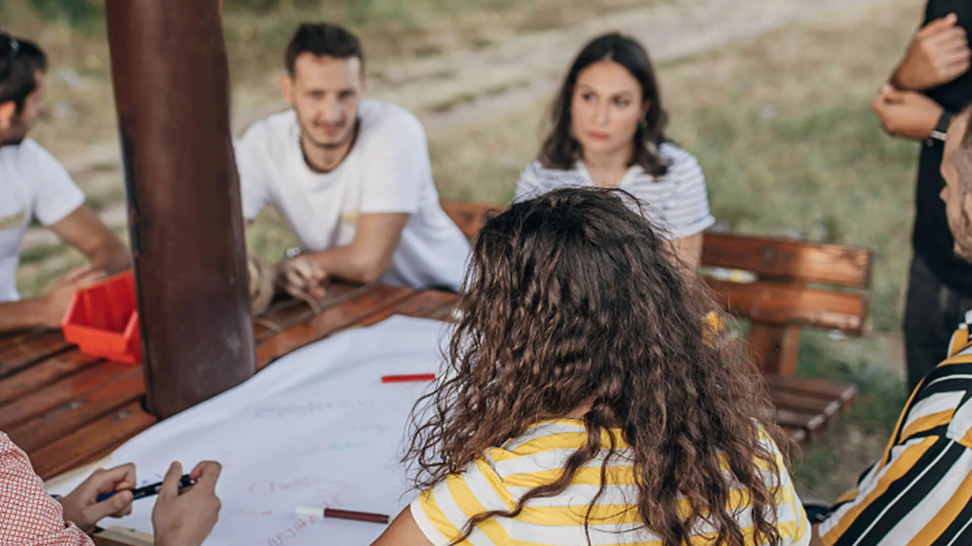 Group of young adults sitting around a wooden table outdoors, discussing and writing on a large sheet of paper.