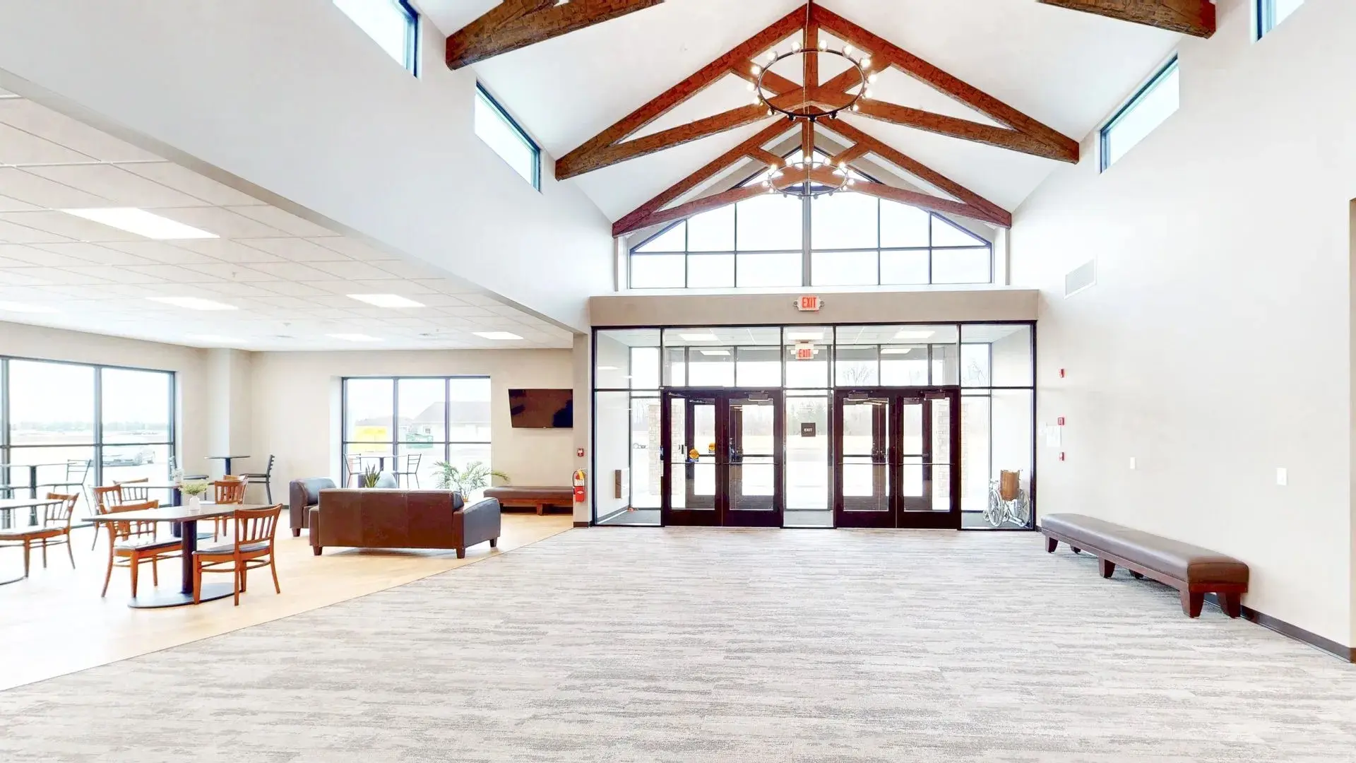 Spacious church lobby with high ceiling, wooden beams, glass double doors, seating area with tables and chairs, and large windows.