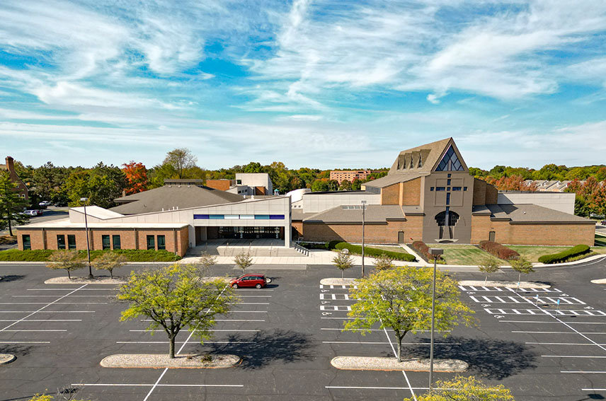 Modern church building with large parking lot and a few trees under a partly cloudy blue sky.