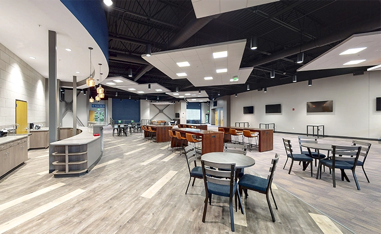 Modern cafe interior with gray wood flooring, round tables with chairs, bar seating with orange stools, and multiple flat-screen TVs on white walls.