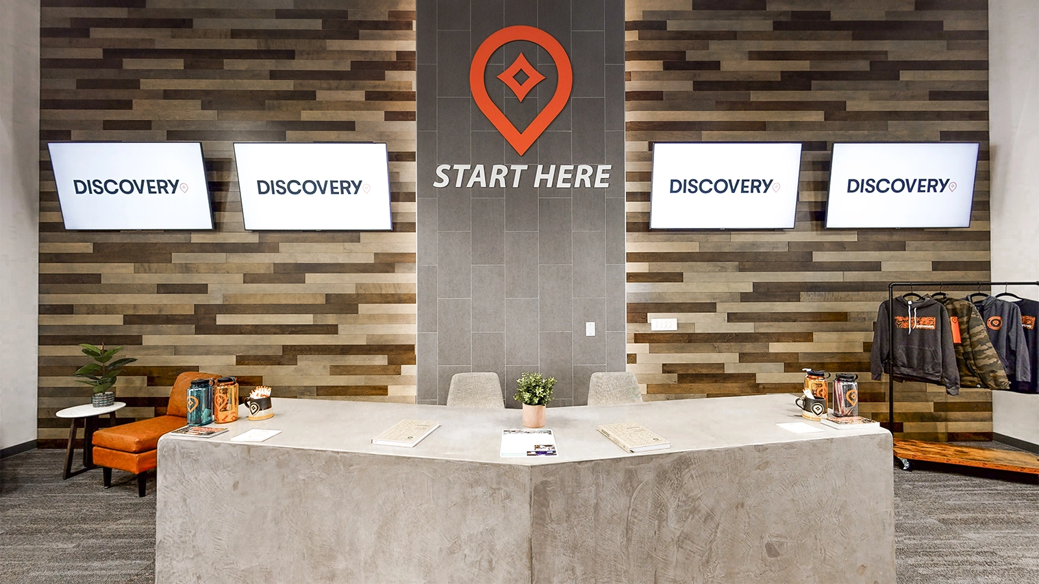 Modern reception area with a concrete desk, wooden wall panels, four screens displaying 'DISCOVERY,' and 'START HERE' text with a location icon above the desk.