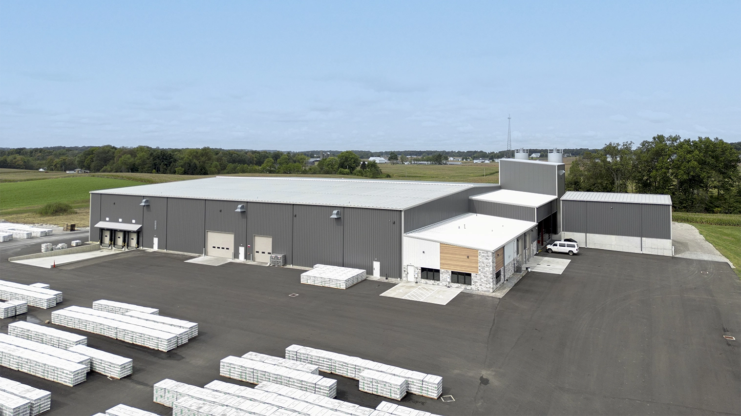A large industrial warehouse with a gray exterior, loading docks, and stacks of packaged materials in the parking lot.