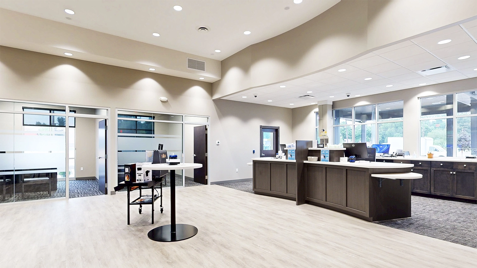 Modern bank interior with wooden counters, large windows, glass office doors, and a round table with promotional materials.