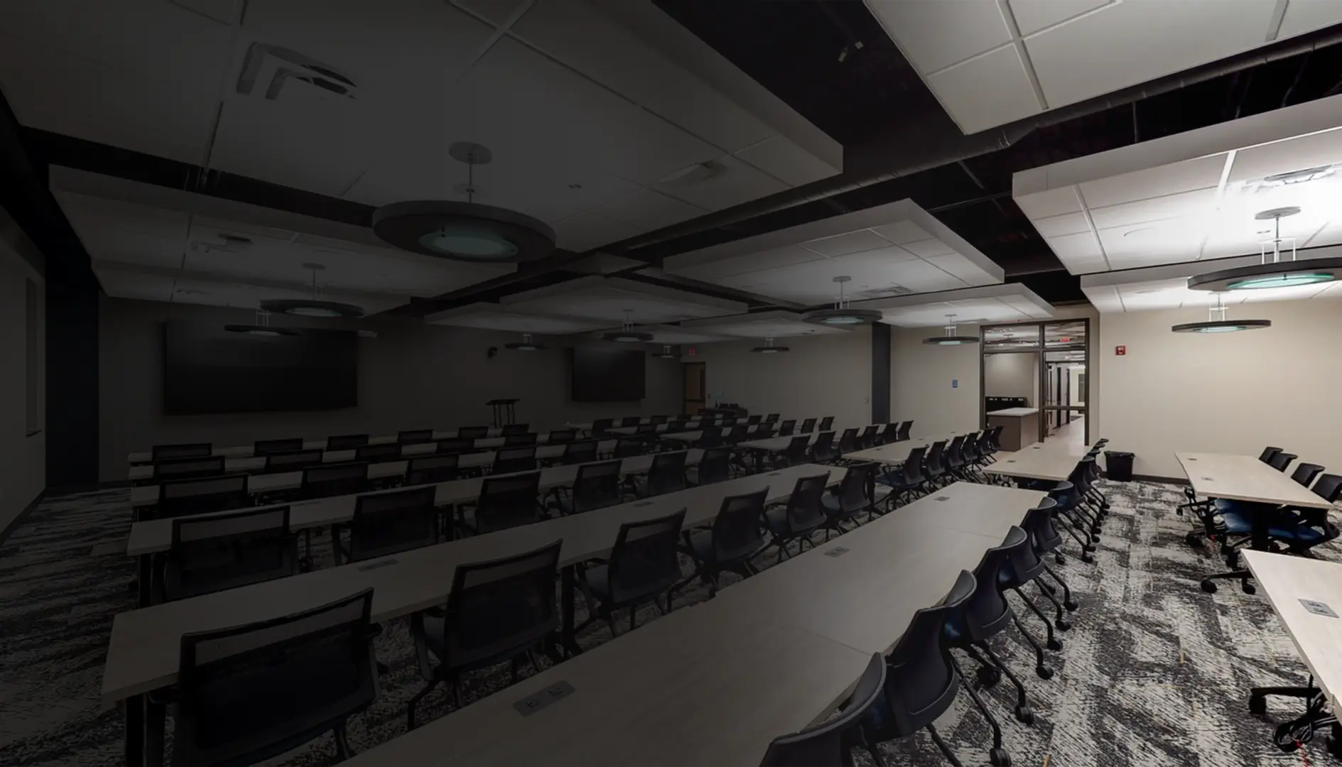 Large modern conference room with rows of tables and black chairs on patterned carpet and ceiling lights.