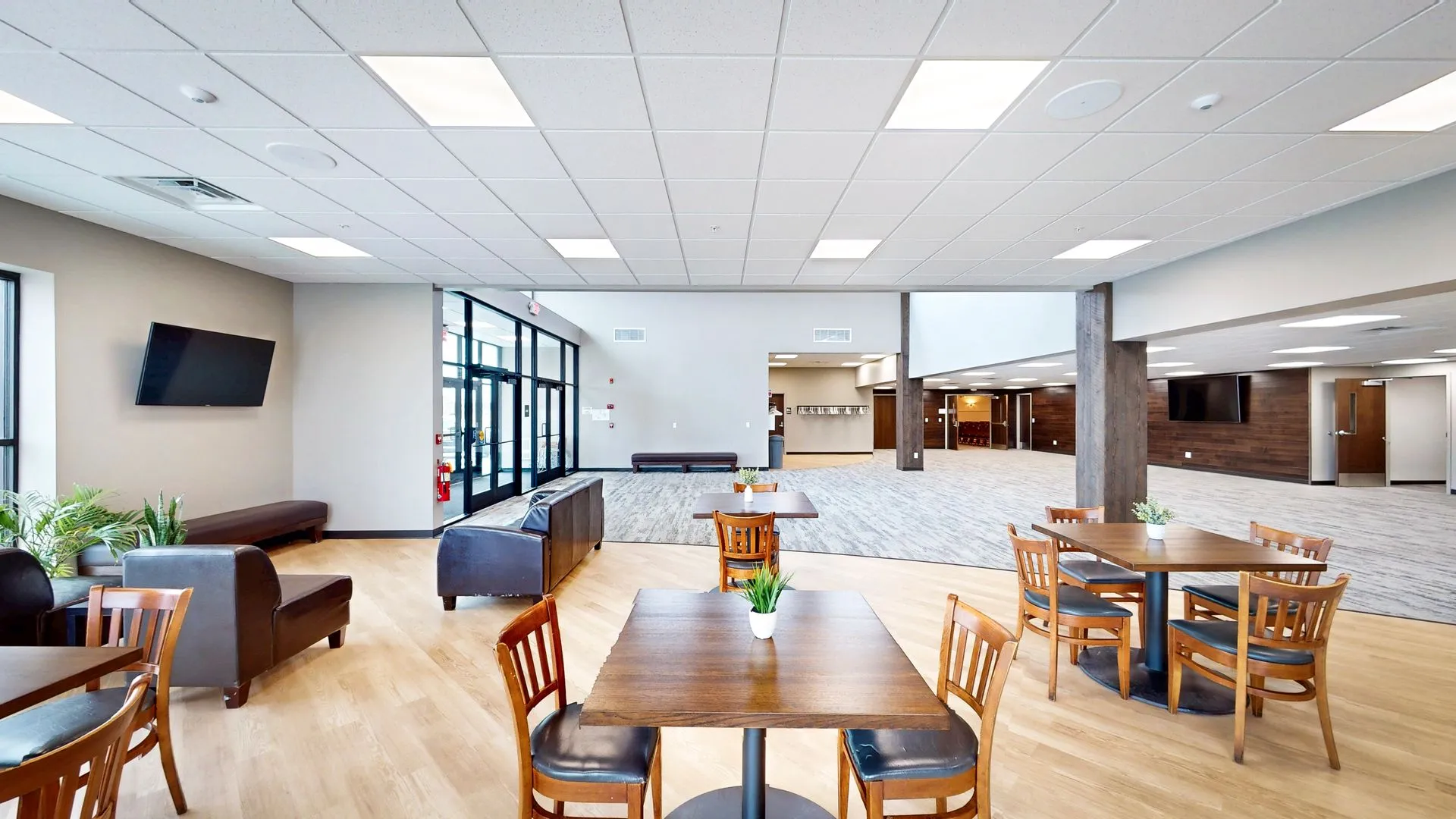 Bright modern lobby with light wood flooring and gray carpet, featuring wooden tables with chairs and small plants, leather armchairs, a bench, wall-mounted TVs, large windows, and glass entrance doors.