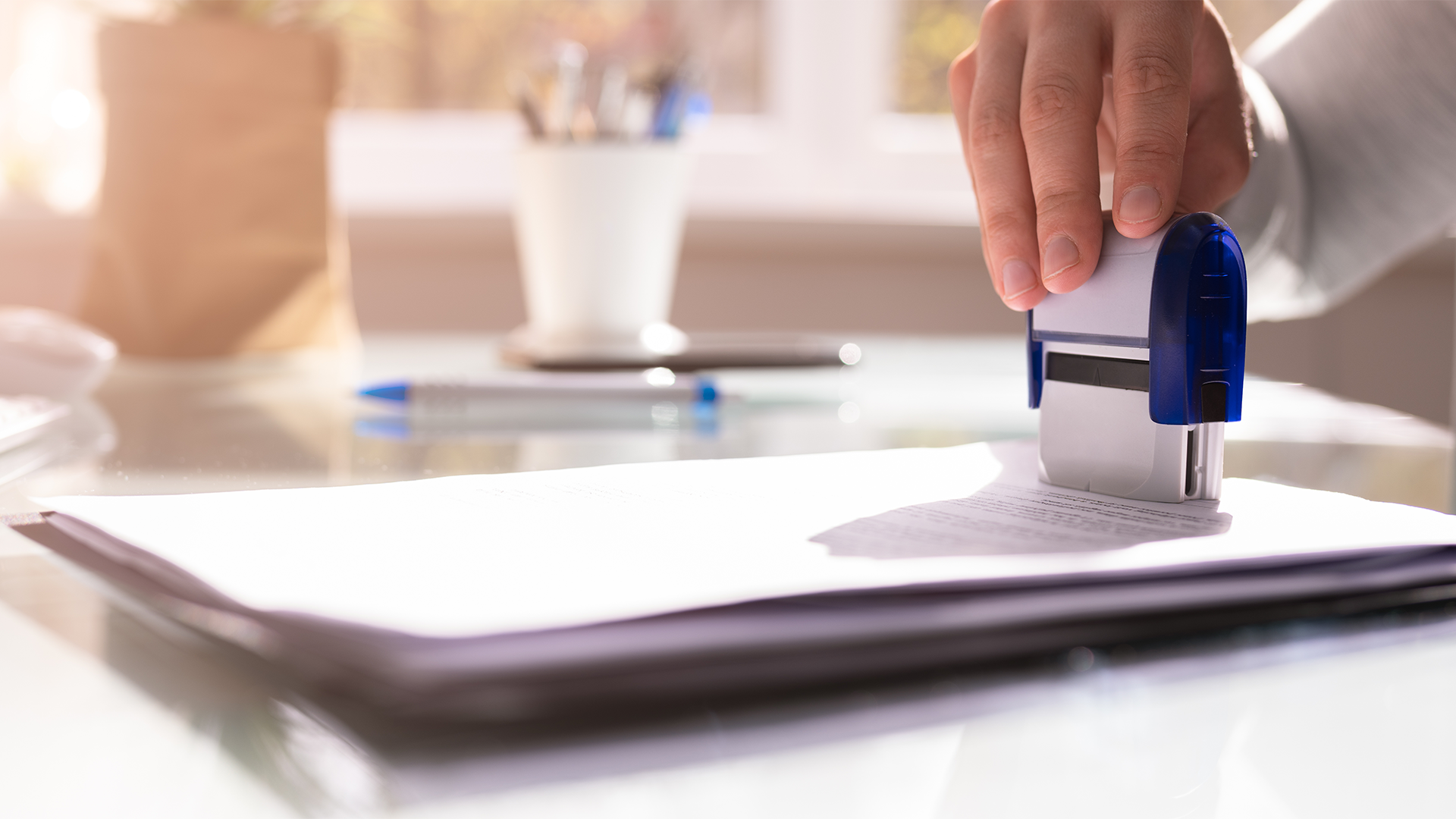 Close-up of a hand pressing a blue and white rubber stamp onto a stack of documents on a glass desk with a blurred pen and cup in the background.