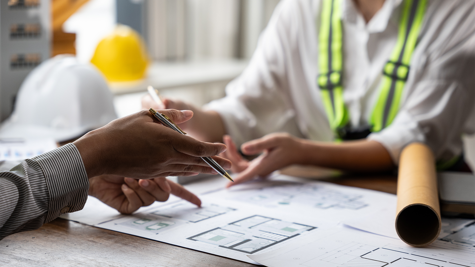 Two people examining architectural floor plans on a wooden table, one holding a pen and pointing, with construction helmets and a rolled blueprint nearby.
