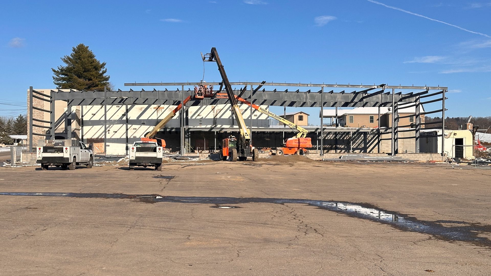 Construction site with metal frame structure of a building being assembled on a clear day, with two white utility trucks and three orange cranes lifting workers and materials.