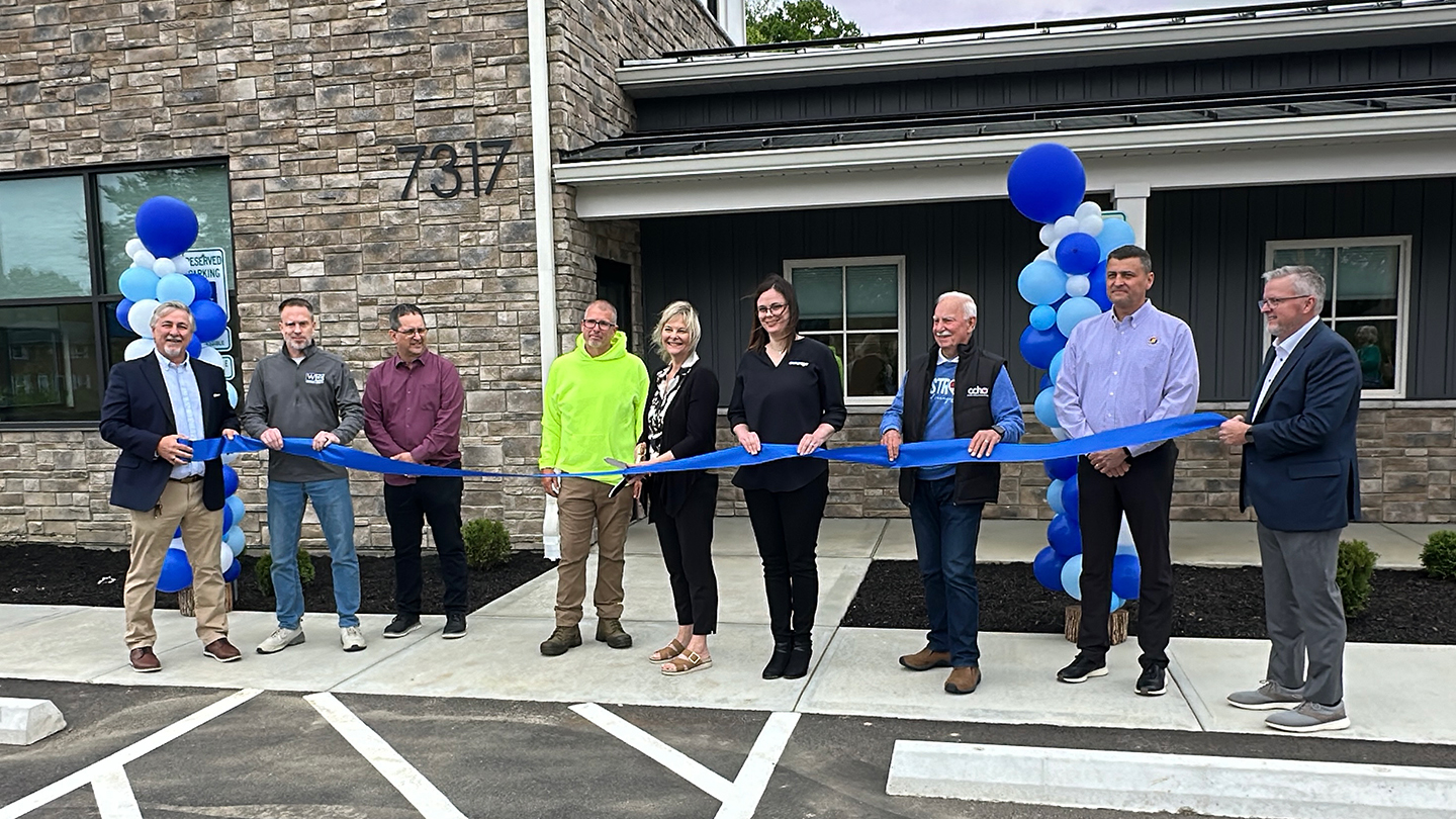 Nine people standing in front of a stone building at address 7317, holding a long blue ribbon for a ribbon-cutting ceremony, flanked by blue and white balloon columns.