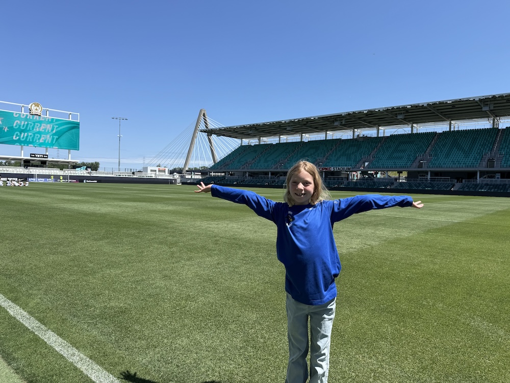 A child wearing a blue shirt stands on a soccer field with outstretched arms on a sunny day