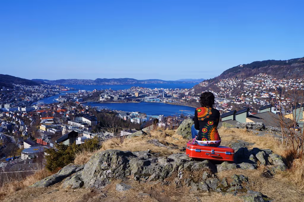 Person sitter på en rød koffert på en høyde med utsikt over en by ved vannet under en klar blå himmel.