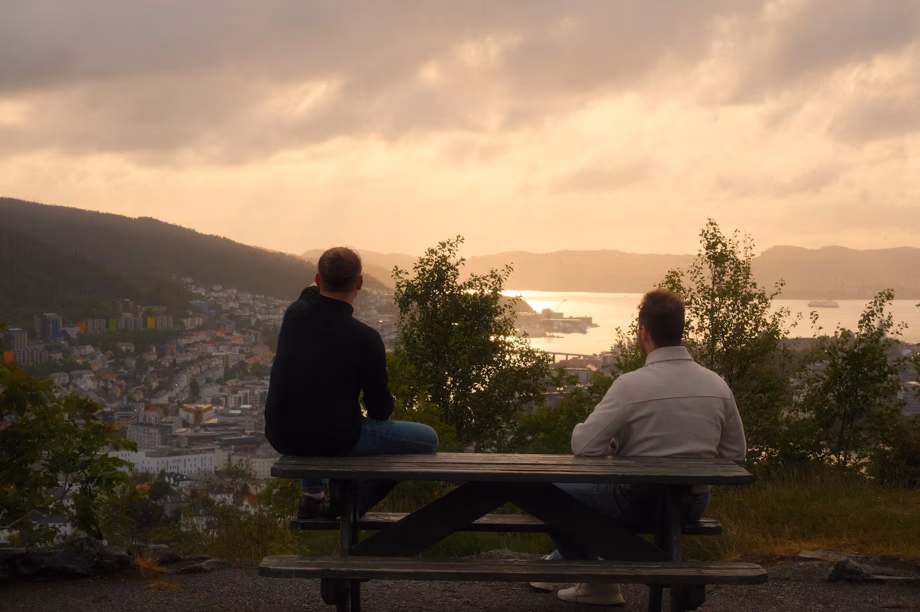 Two people sitting on a bench looking out over a city and a body of water under a sunset sky.